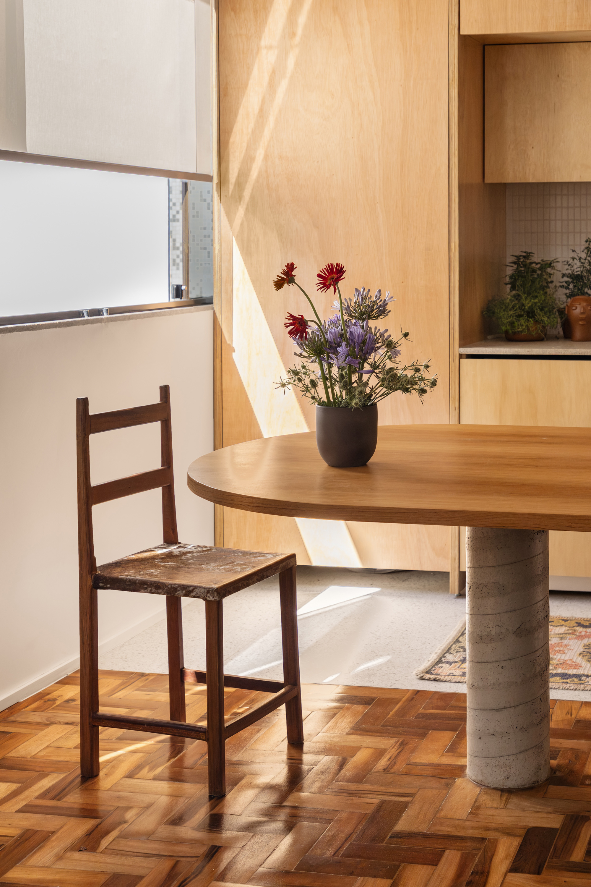Wooden ladder-back chair and flower vase on the oak table, with afternoon sun striking the plywood cabinet wall