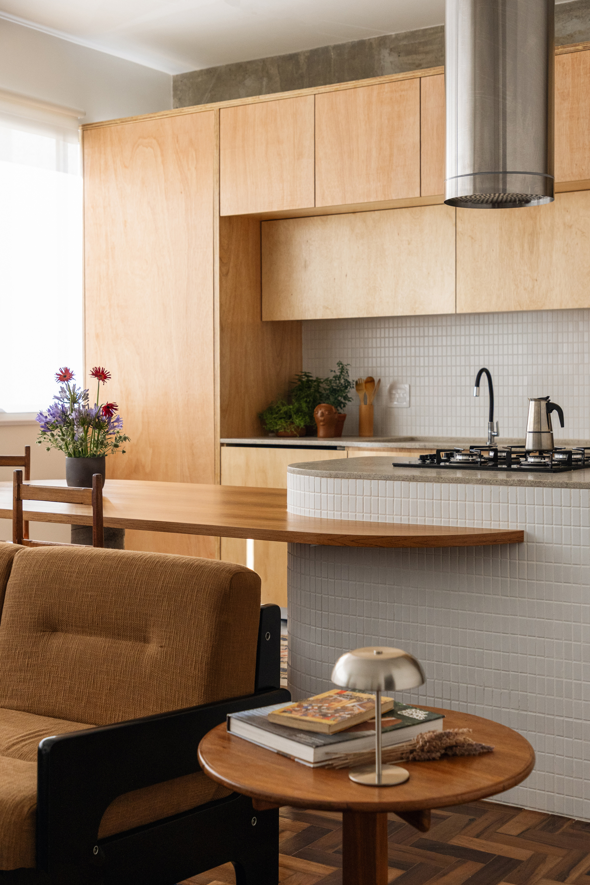 Tile-clad island and plywood cabinets seen past an armchair and round side table with books and a lamp