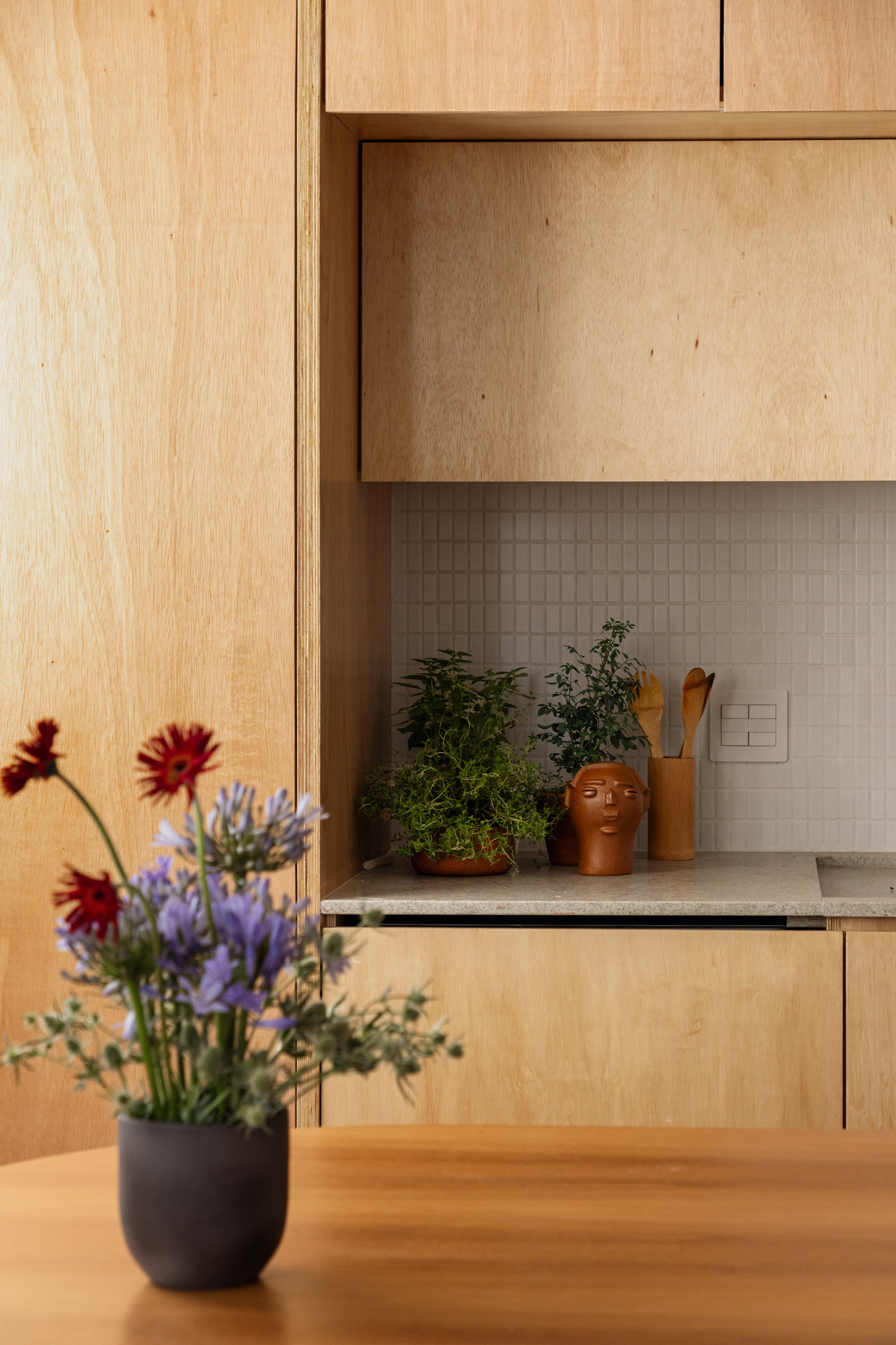 Plywood kitchen cabinets above a white mosaic tile backsplash with potted herbs, a terracotta face vase and a bouquet in the foreground