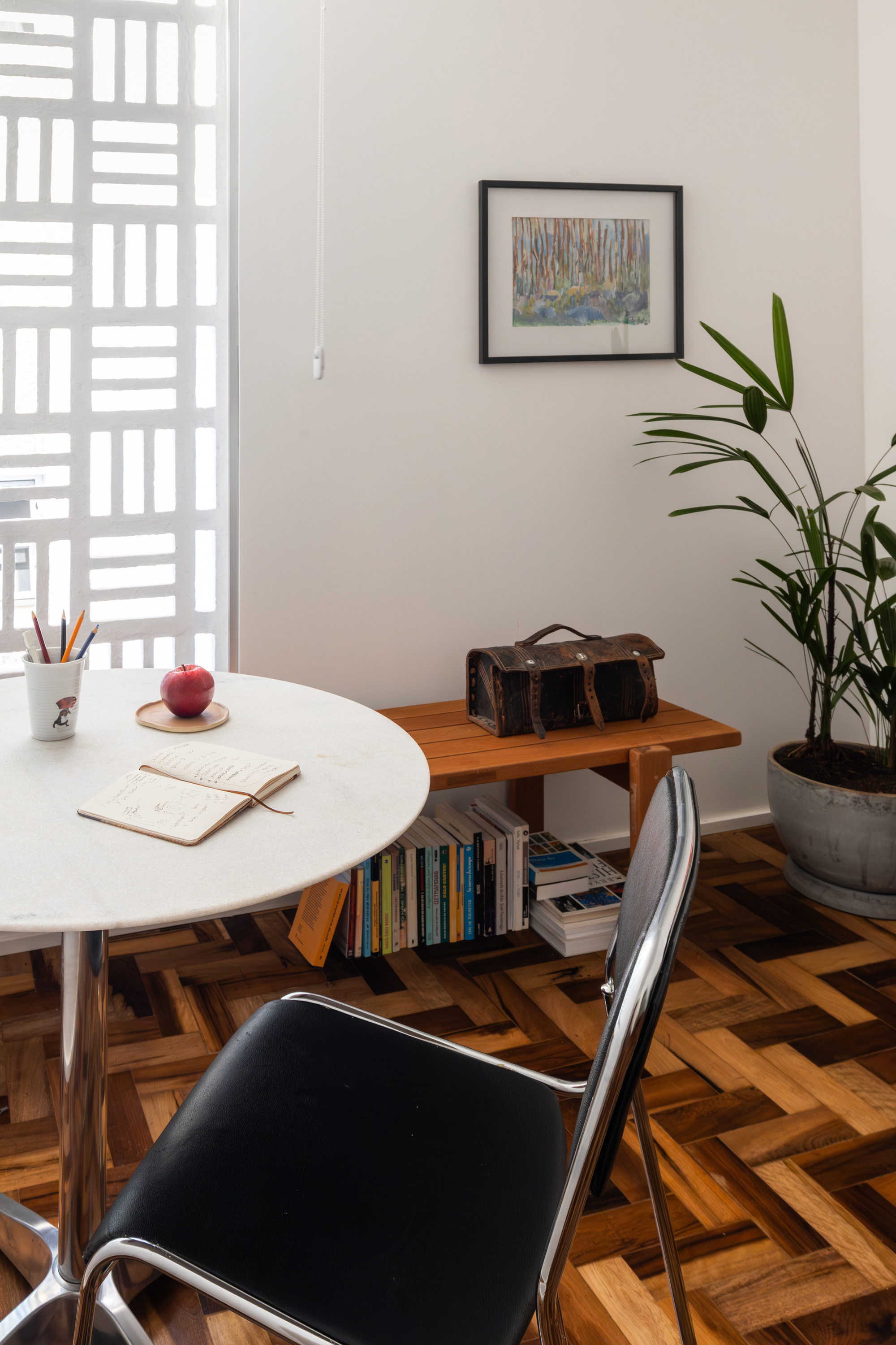 Round marble-top table with a chrome and black chair beside a low wooden bench, books and a plant on herringbone floor