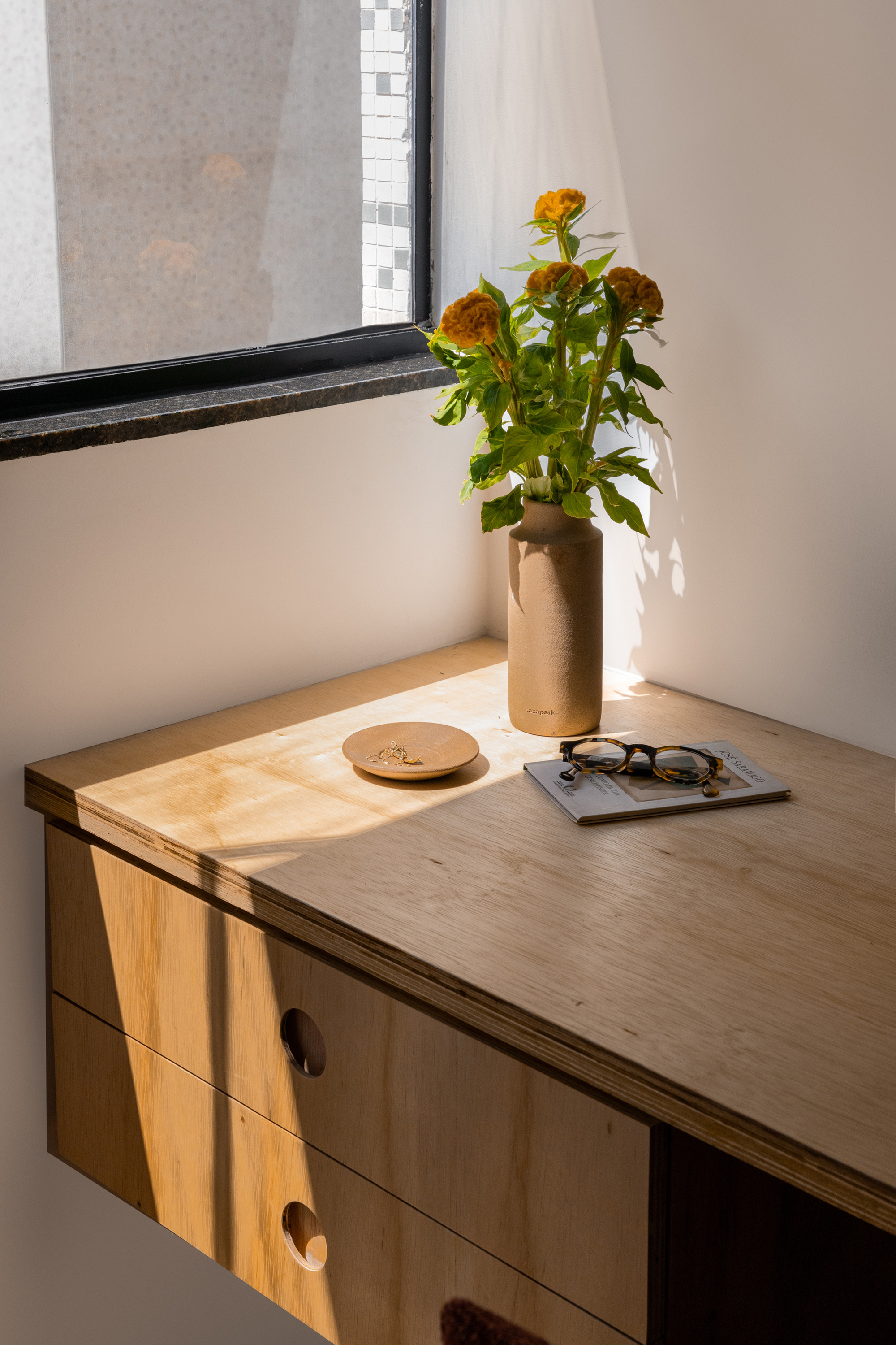 Wall-mounted plywood desk with round finger-pull drawers, a vase of orange flowers and glasses in a patch of afternoon sun