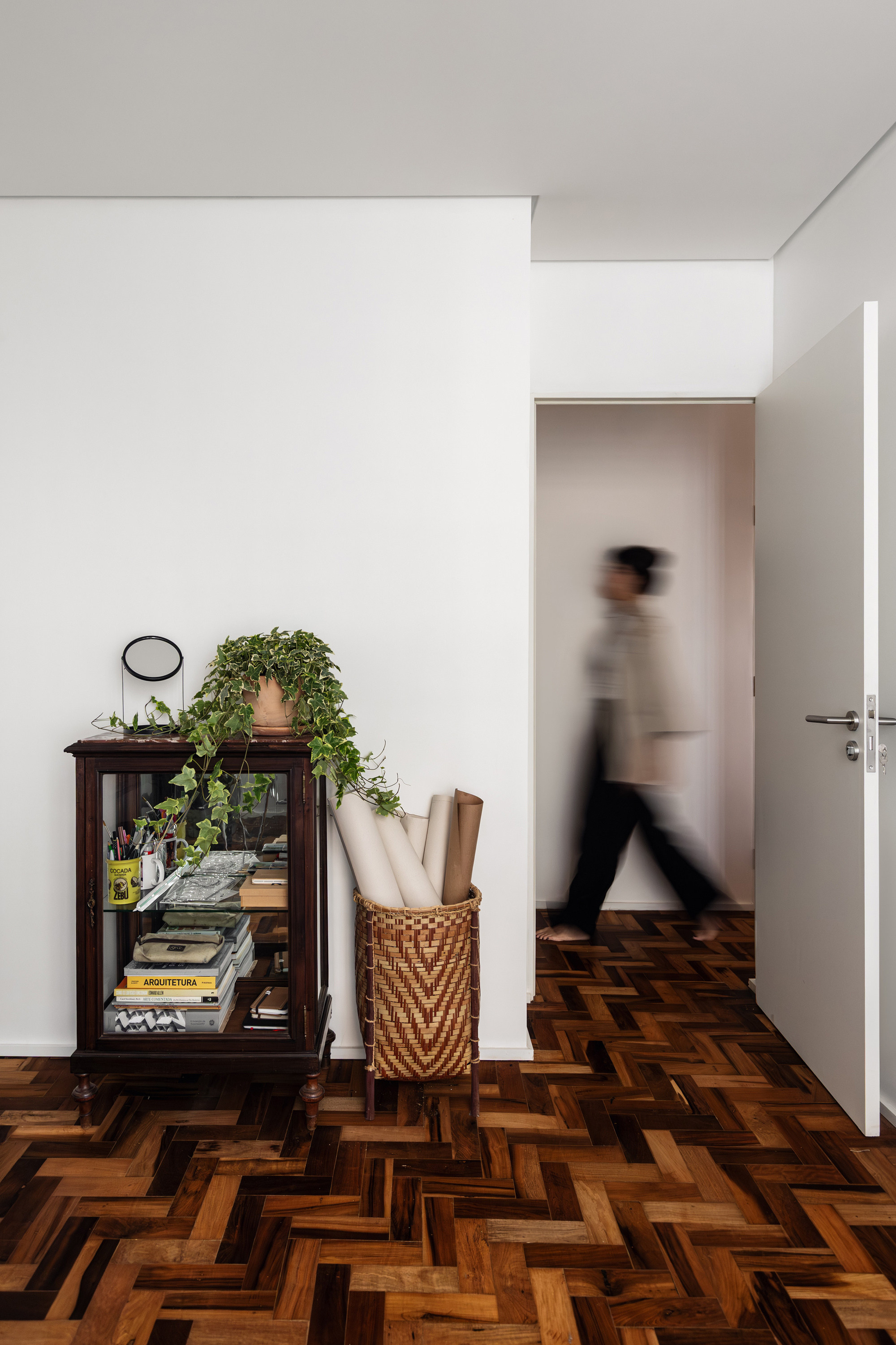 Glass-front wooden cabinet with trailing ivy beside a woven basket of rolled papers, as a blurred figure walks through the doorway