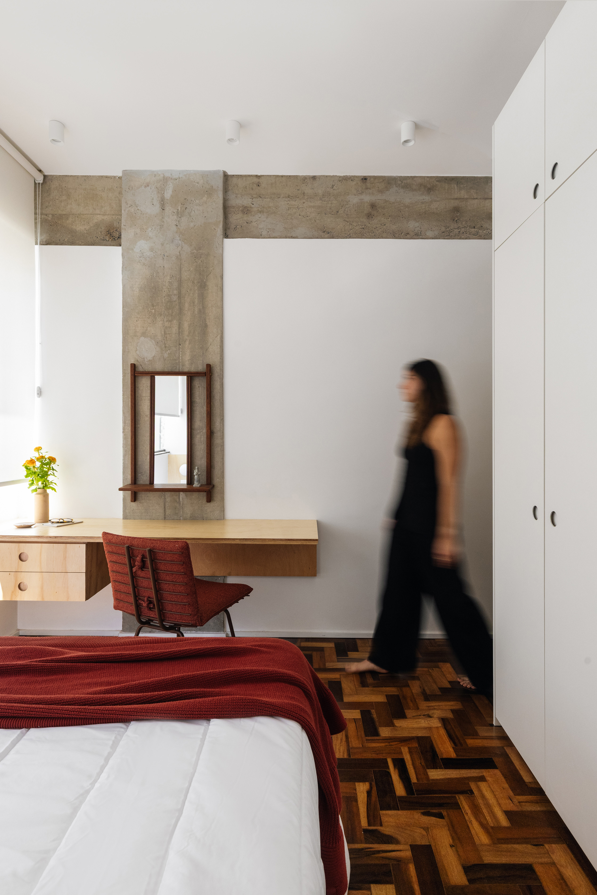 Bedroom corner with an exposed concrete column, floating plywood desk and red chair, as a blurred woman walks past white wardrobes