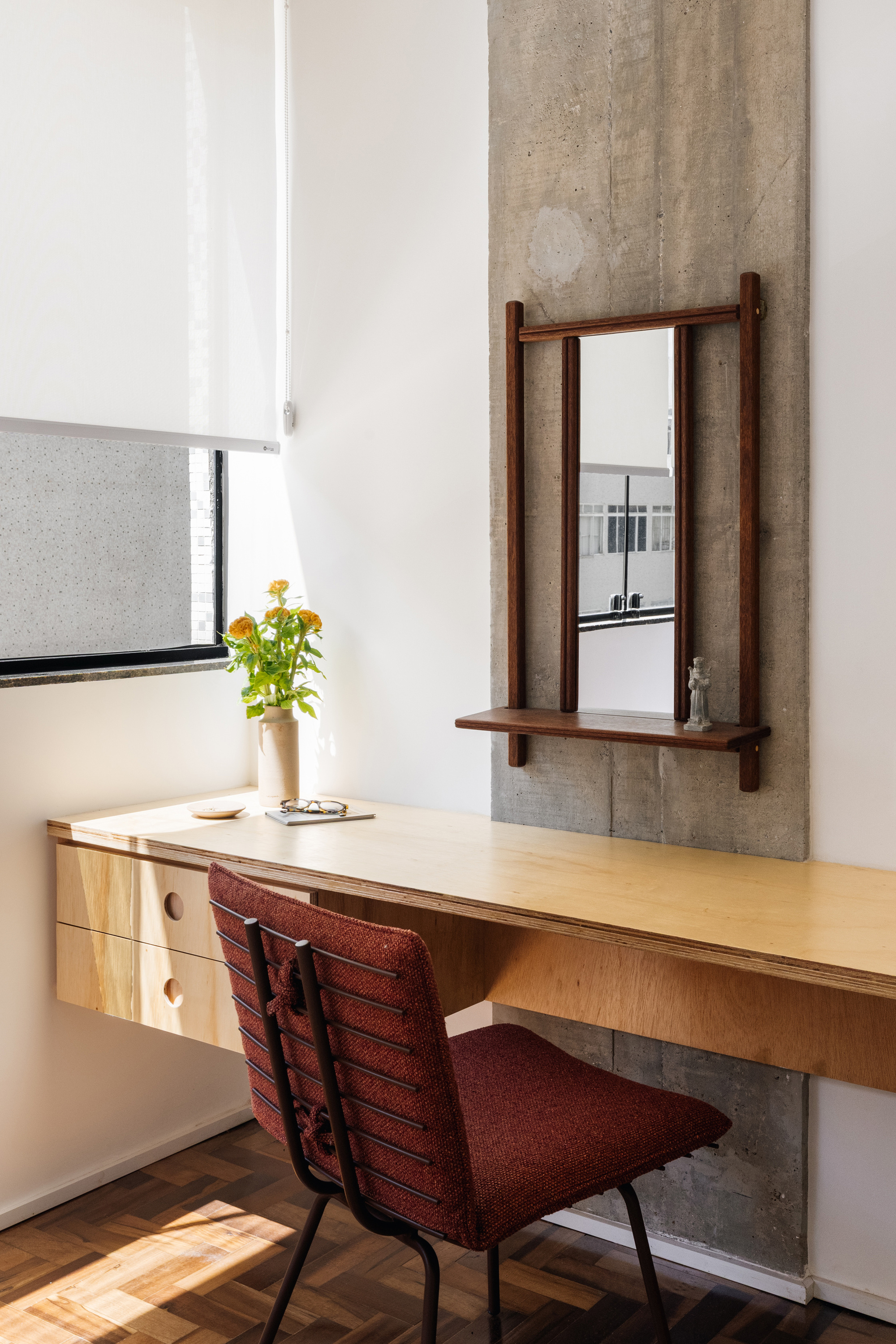 Plywood desk cantilevered from the wall beside a board-marked concrete strip with a timber-framed mirror, red chair and flowers in afternoon sun