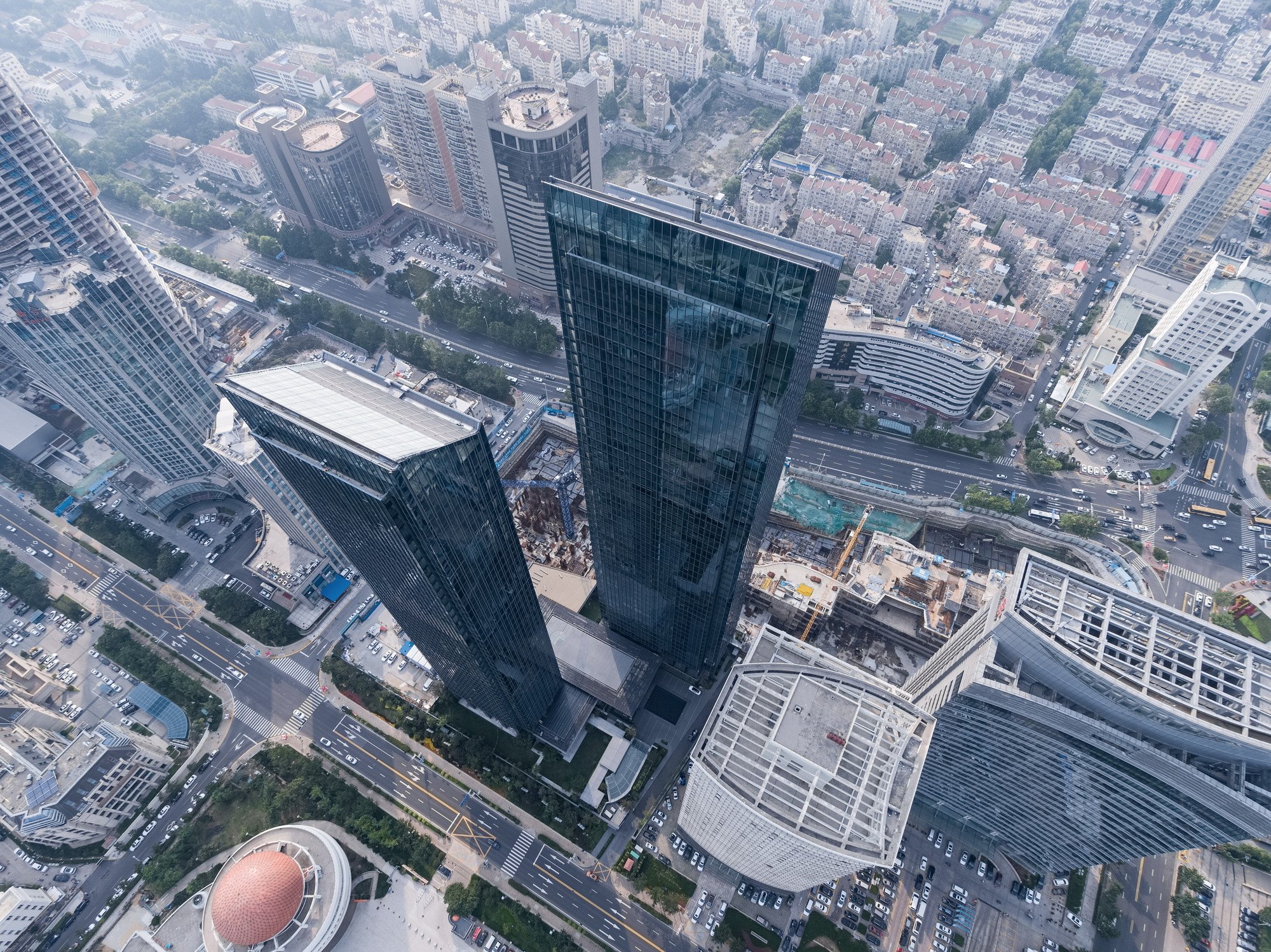 Top-down drone view of glass towers, a construction site and surrounding streets in hazy daylight