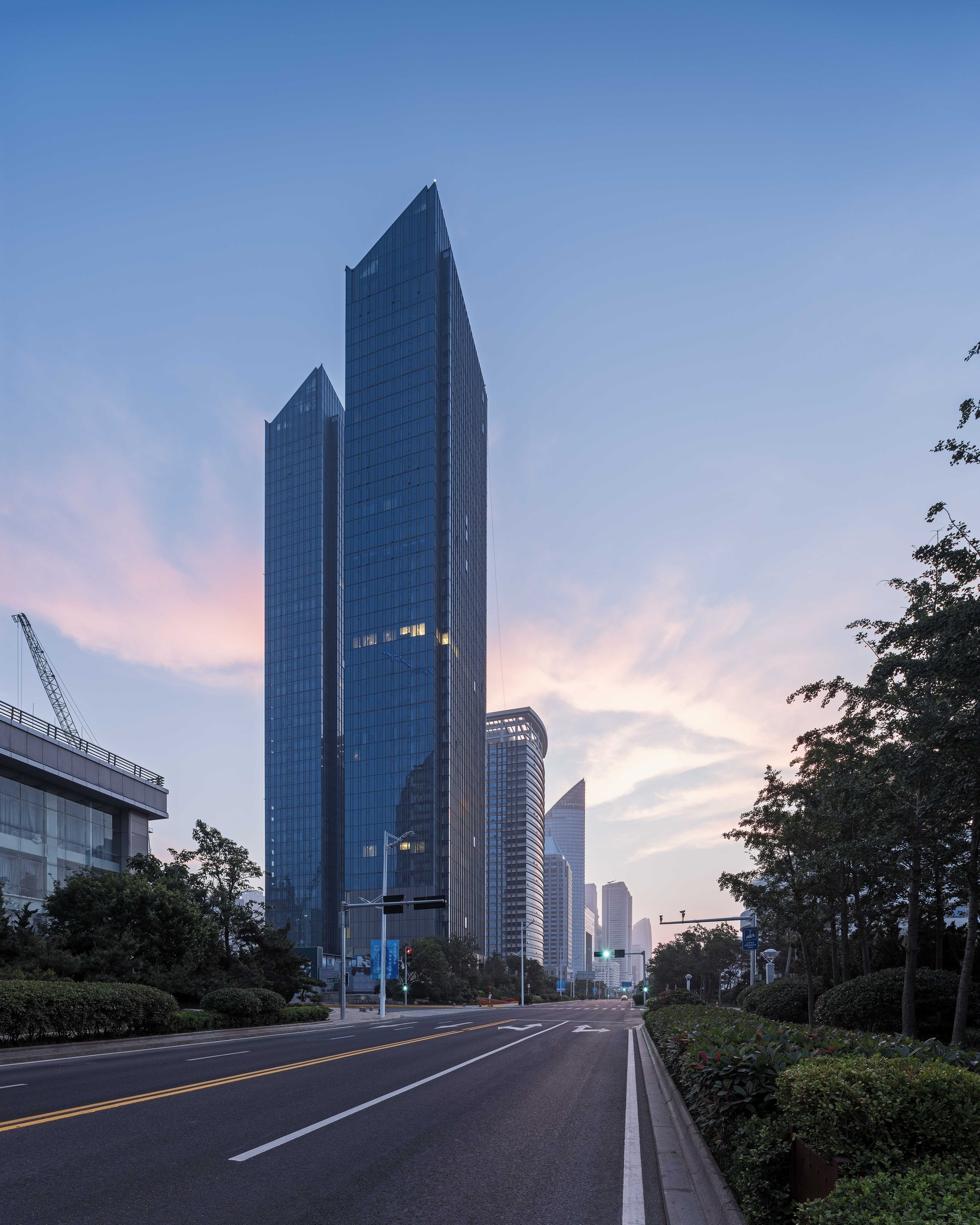 Street-level view of two glass towers with angled tops beside a tree-lined road at dusk