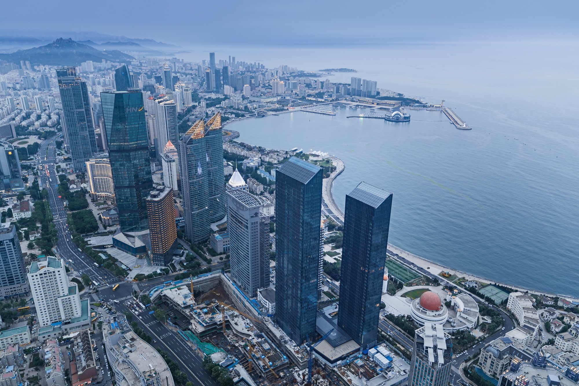 Aerial view of twin glass towers above a curving shoreline and bay under a misty sky