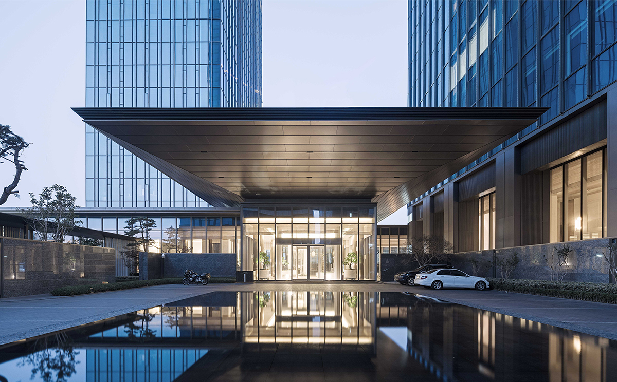 Entrance canopy cantilevered over a glazed lobby, reflected in a still pool at dusk