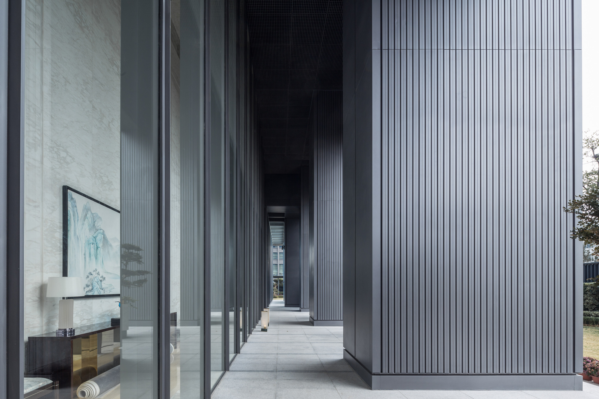 Covered walkway between a glazed lobby wall and vertically ribbed grey metal cladding, with a landscape painting visible inside