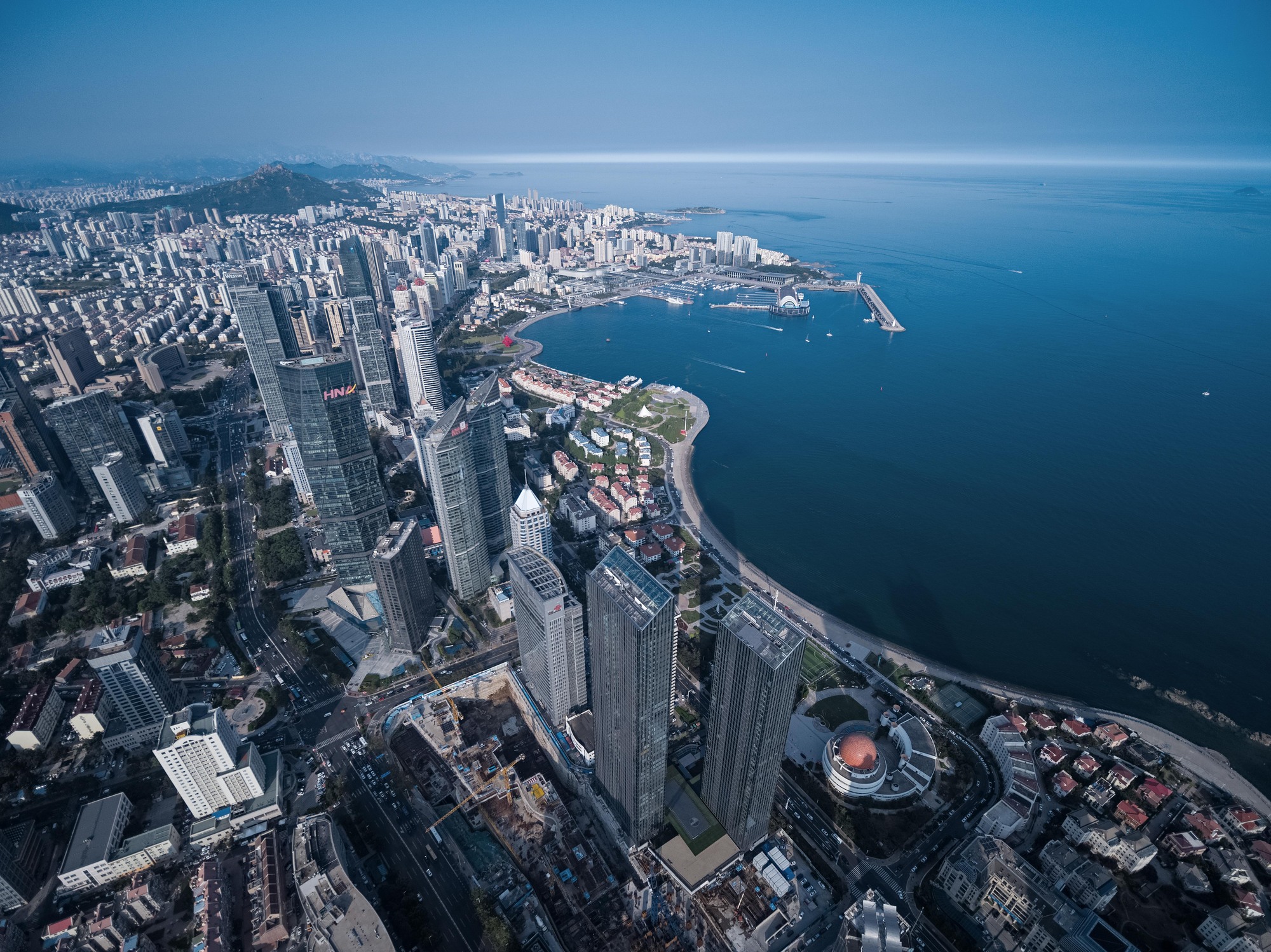 Aerial view of glass towers rising along a curving shoreline, with a harbor pier and dense low-rise blocks under clear haze