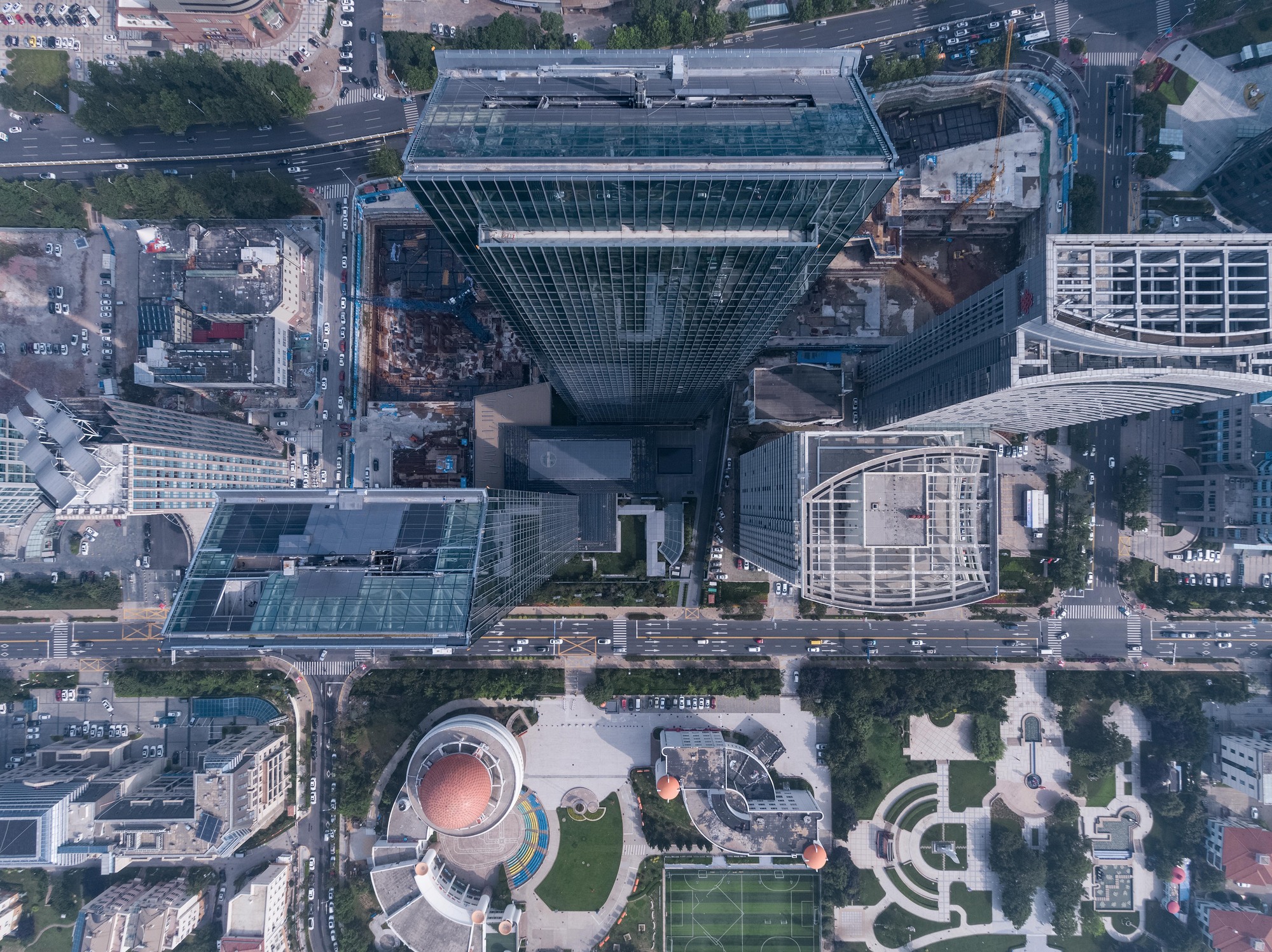 Top-down drone view of a glass tower, neighboring podiums and a landscaped plaza with a sports pitch beside a busy avenue
