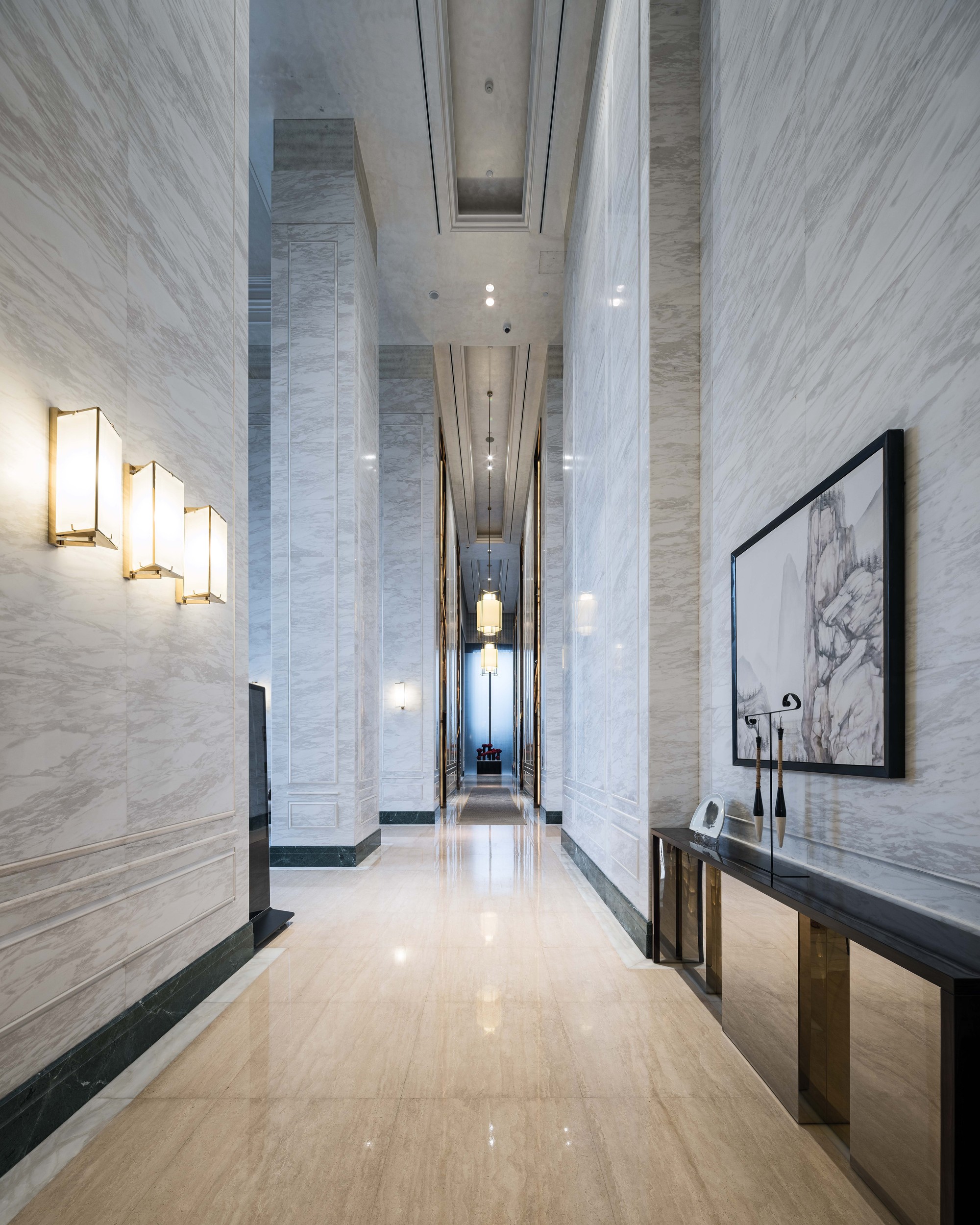 Tall corridor lined with veined white marble panels, brass wall sconces and a framed ink landscape above a console table