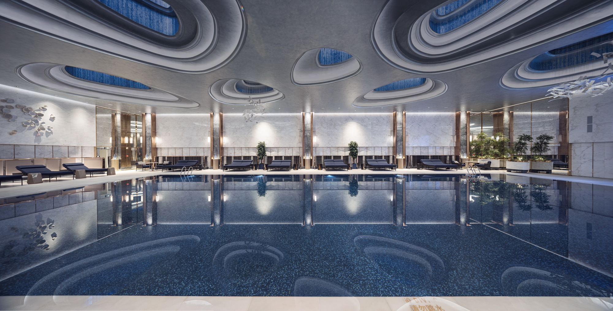 Indoor swimming pool with a ceiling of layered oval skylight rings, loungers along the far wall and potted plants reflected in the water