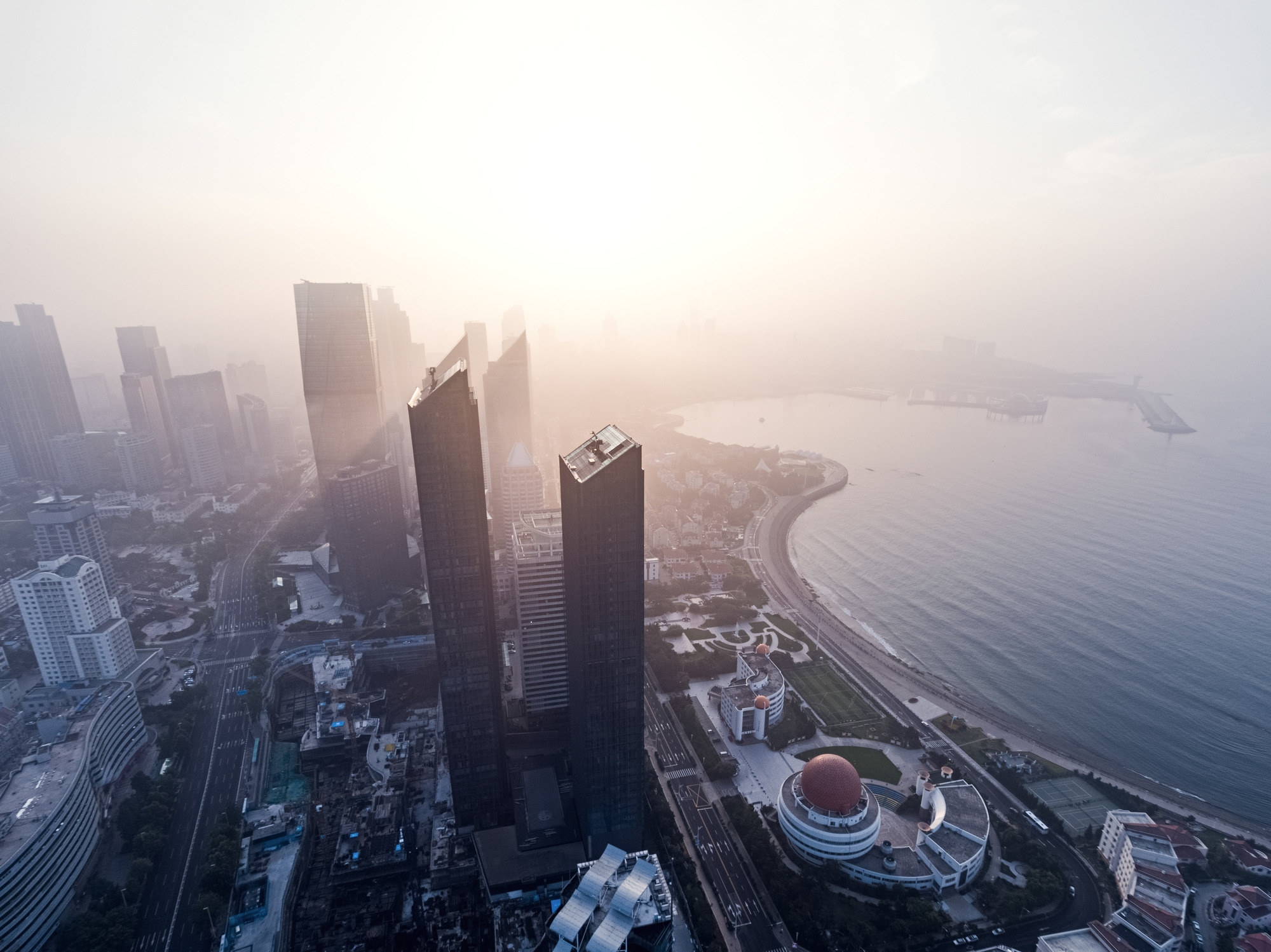 Aerial view of two dark glass towers rising above a curving bay shoreline in hazy morning sun