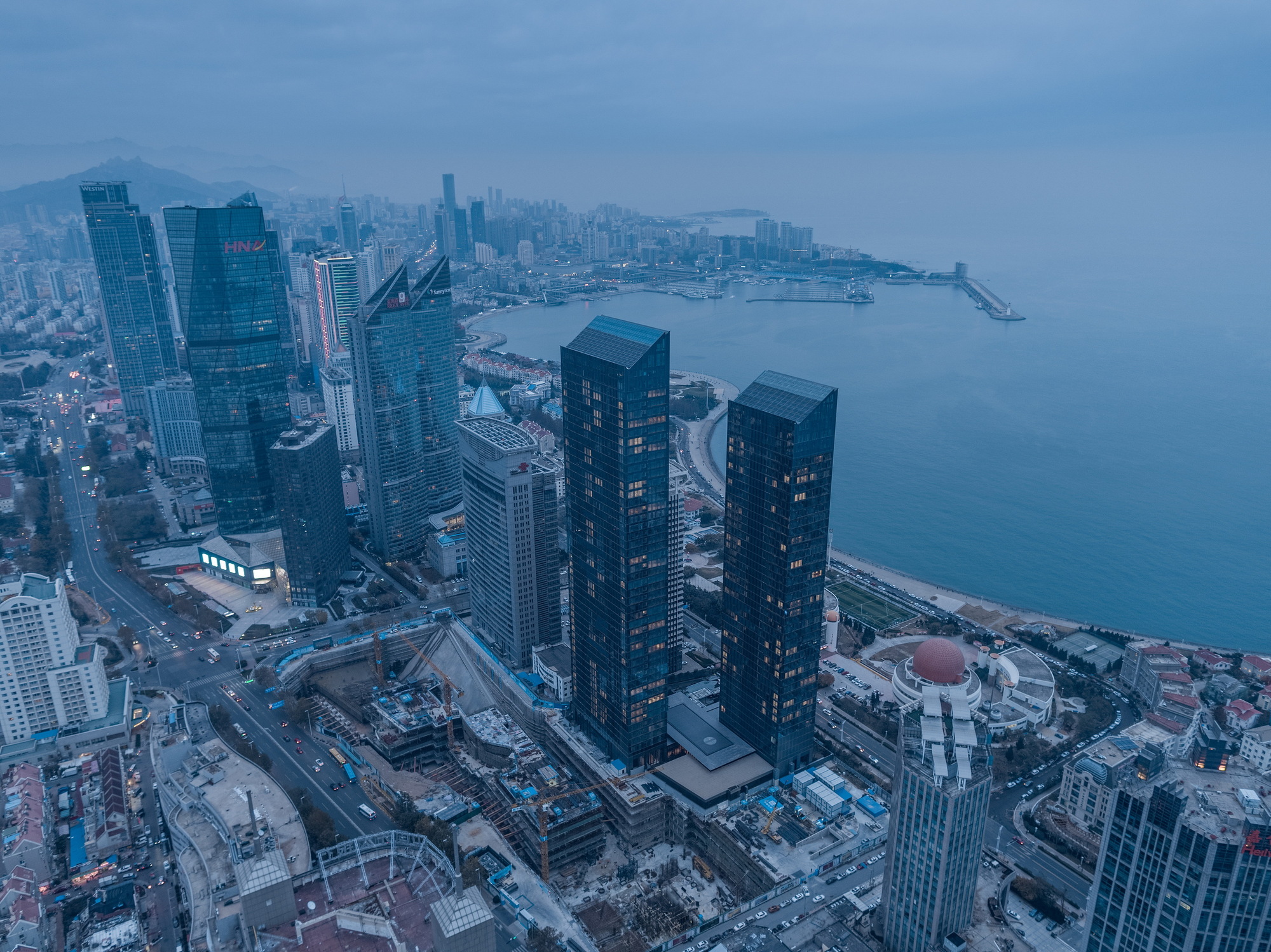 Aerial view of twin glass towers above a waterfront district and harbor pier under a blue dusk sky