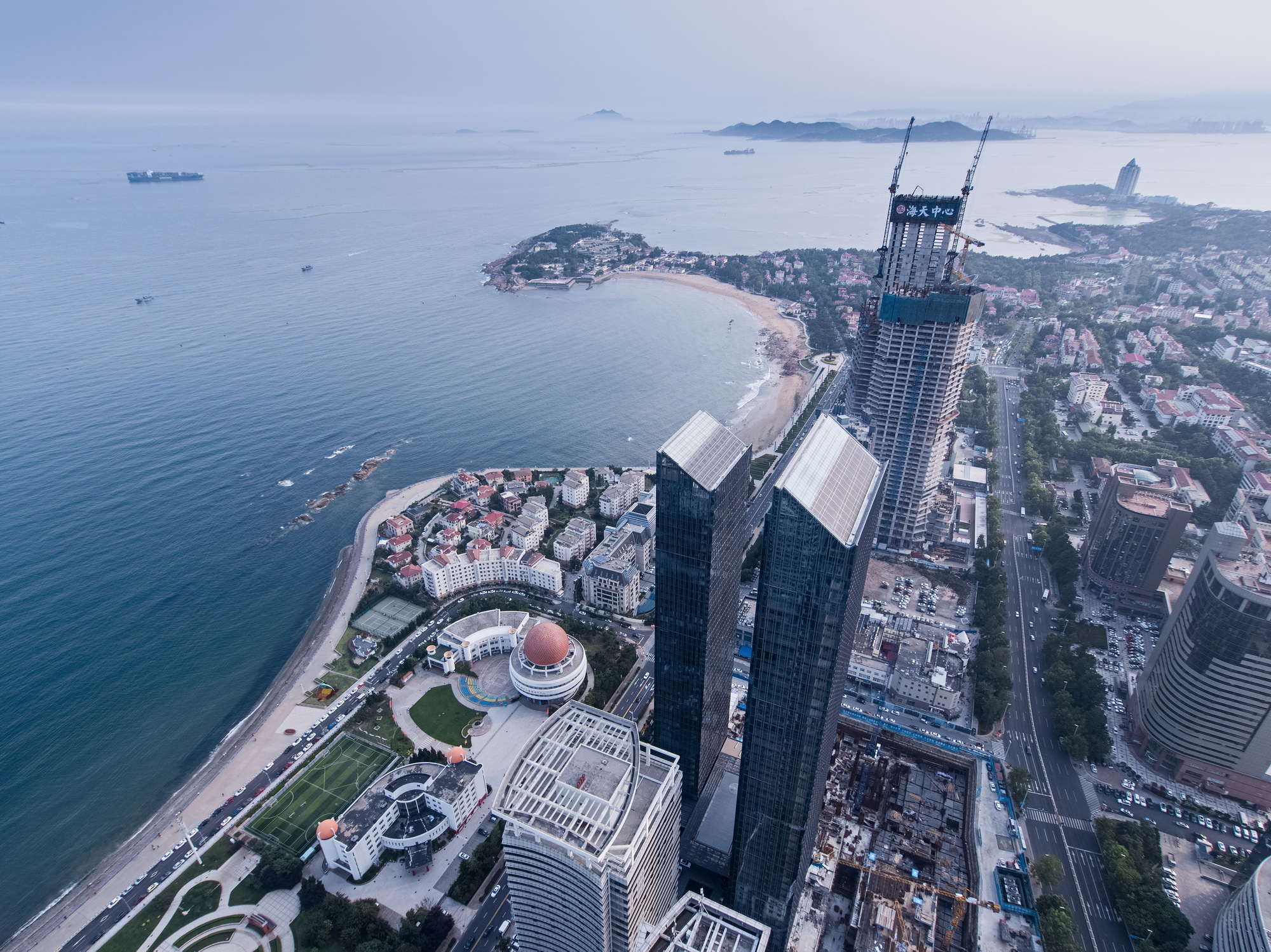 High aerial view of twin glass towers beside a beach cove, with a tower under construction and cranes behind