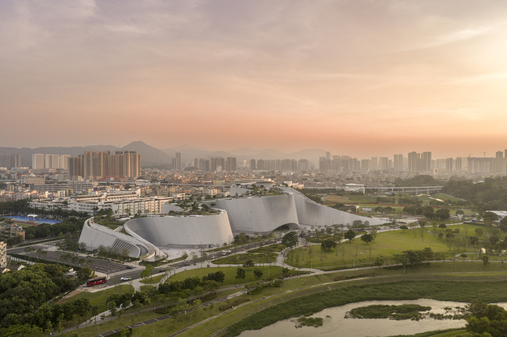 Aerial view of the white ribbed curving volumes beside a river and park, with a hazy city skyline at sunset