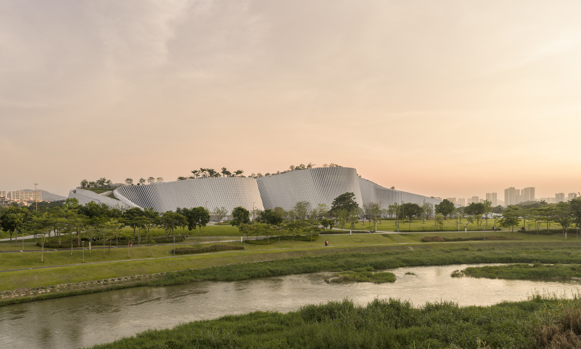 Long view across a river and grassy bank to the fluted white facade with rooftop trees at dusk