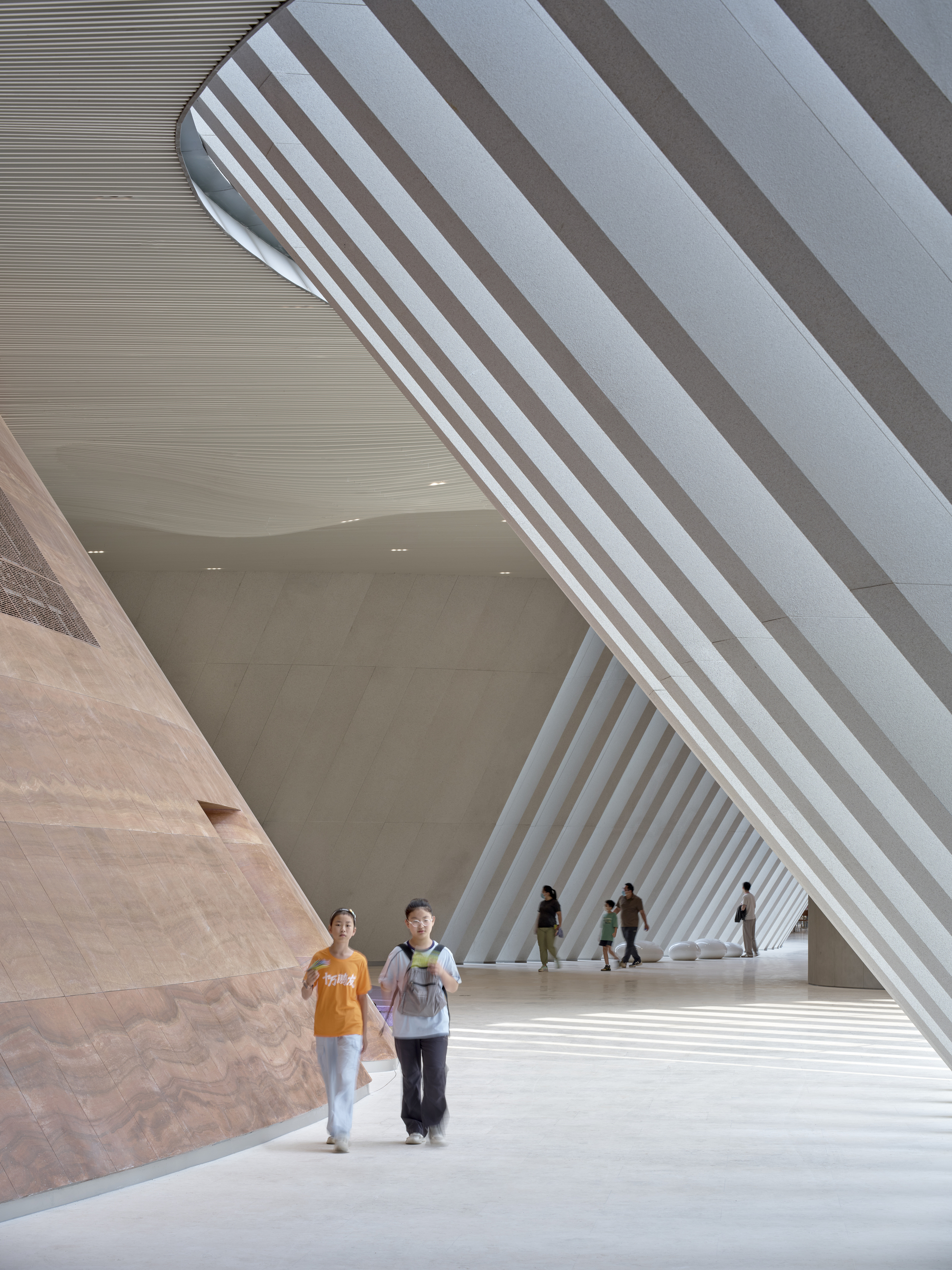 Two visitors walking through a sloping interior passage between a striated stone wall and slanted white fins in daylight