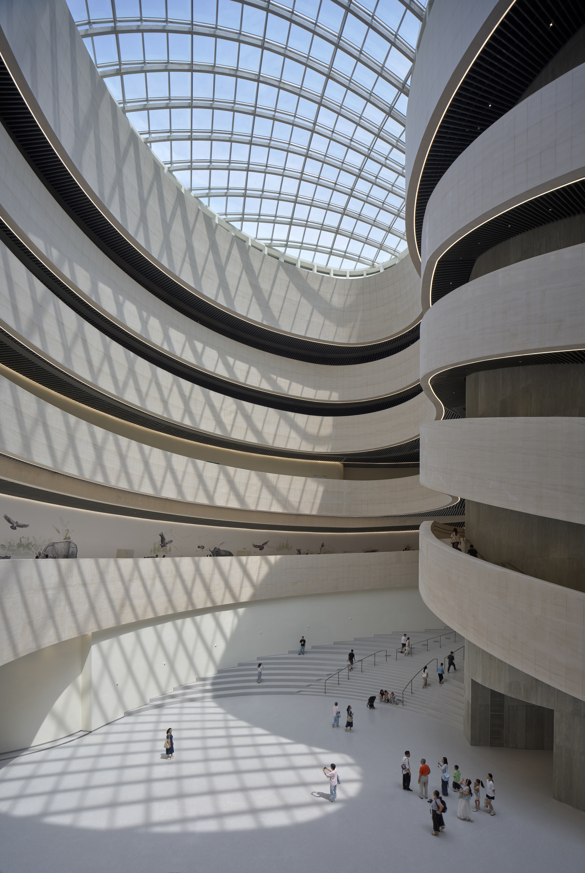 Atrium with stacked curving white balconies under a glass roof, casting lattice shadows on the floor and visitors below