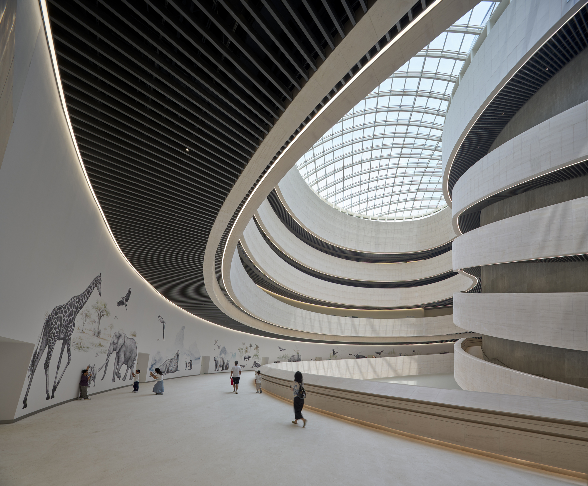 Curved atrium wall painted with a giraffe and elephants, beside a dark slatted ceiling and skylight, with visitors walking