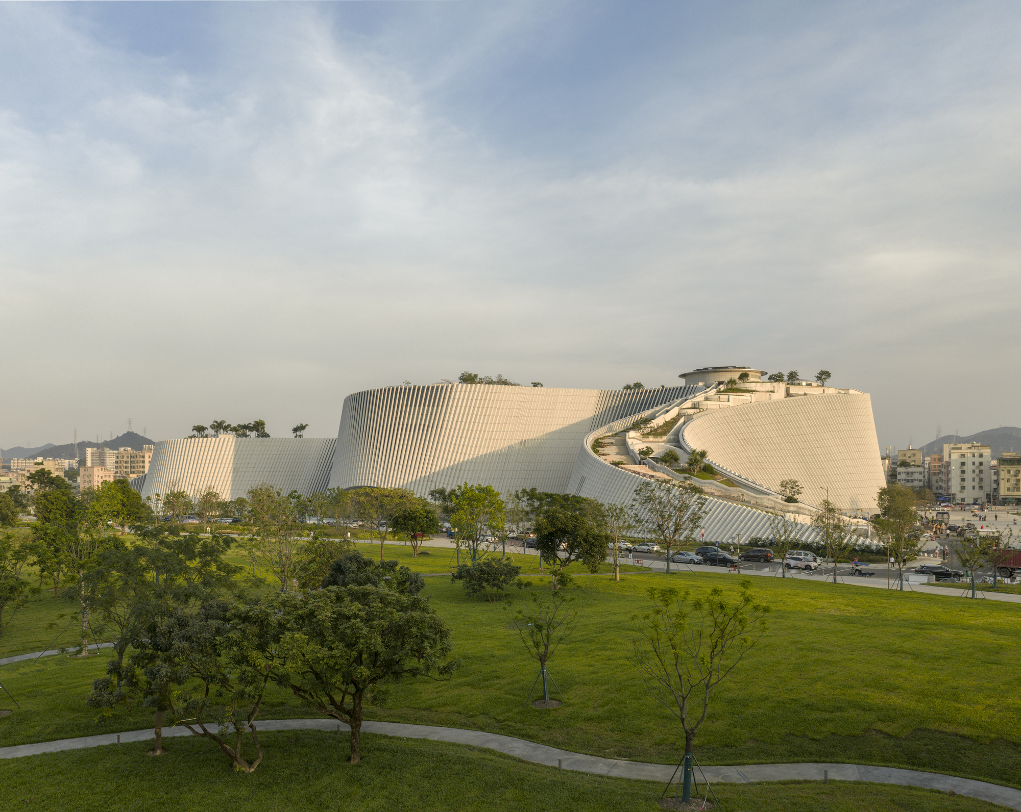 Long view of the white fluted, terraced building mass beyond a lawn with young trees under a hazy evening sky