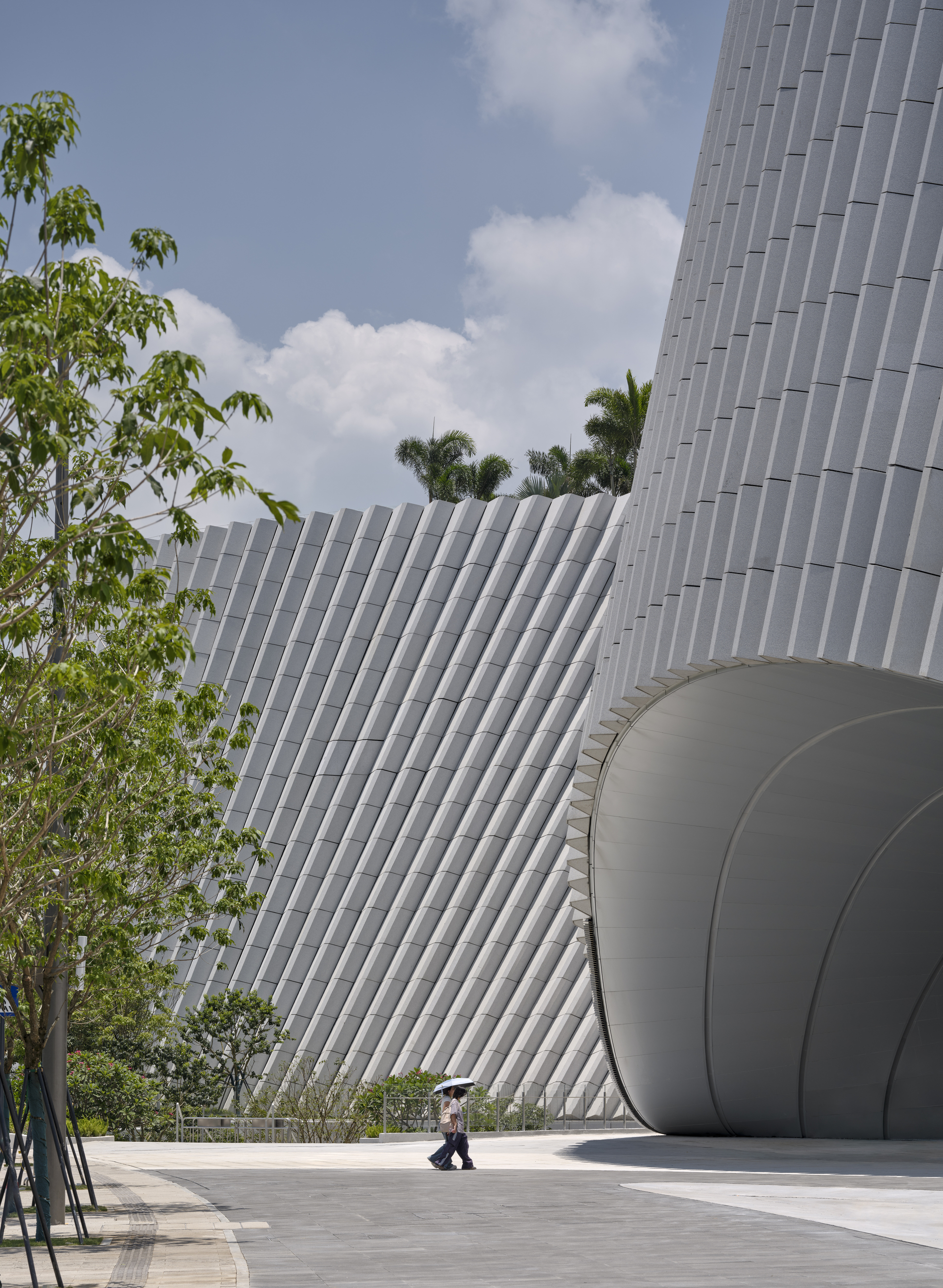 Diagonal white fins on the curved facade beside a rounded entrance opening, with a pedestrian under an umbrella