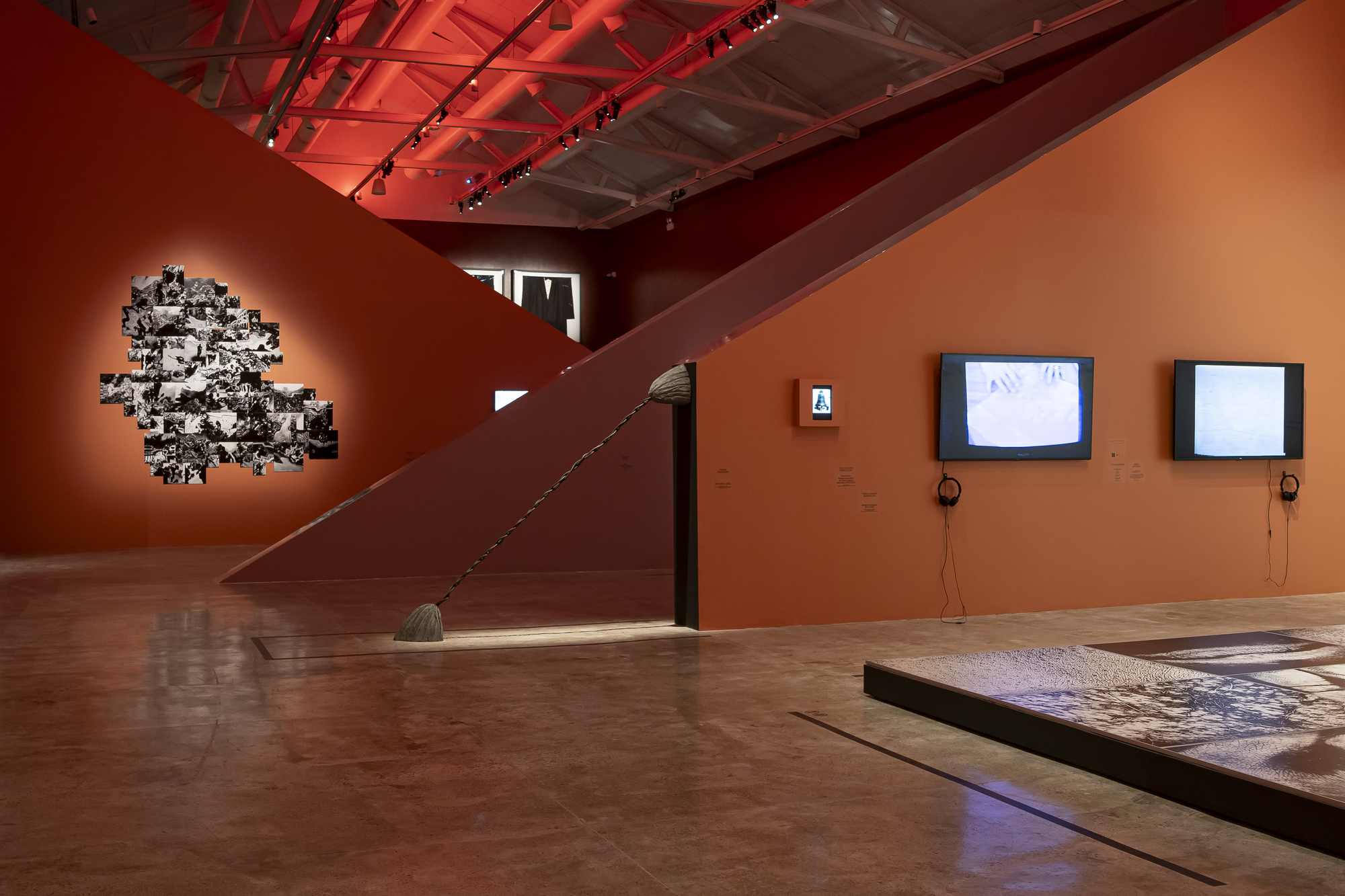 Gallery interior with a slanted orange wall, a hanging broom-like sculpture, a black-and-white photo cluster and two video monitors under a red-lit truss ceilin
