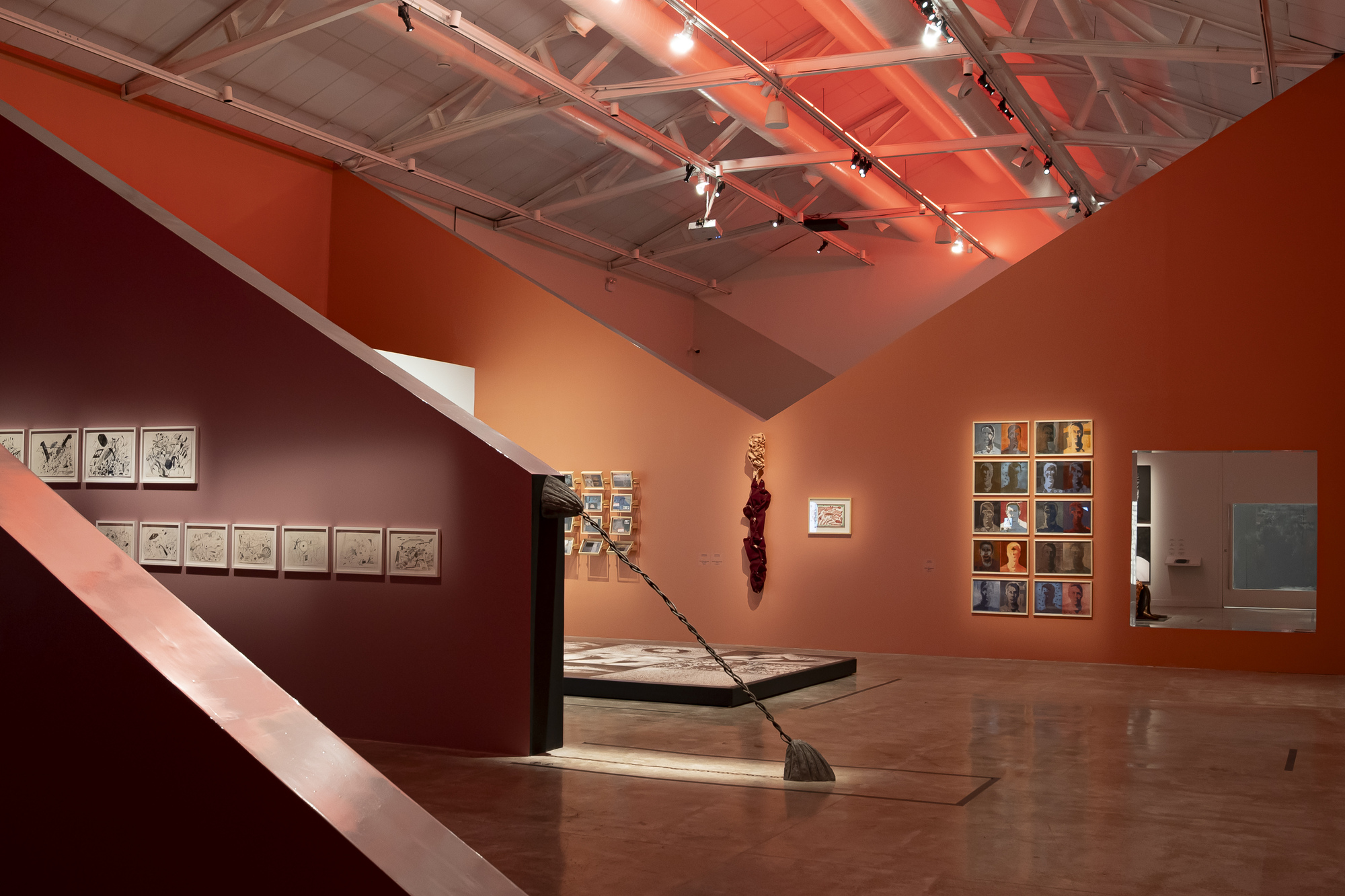 Angled maroon and orange partition walls frame framed drawings, a grid of portraits and a floor platform beneath exposed steel trusses