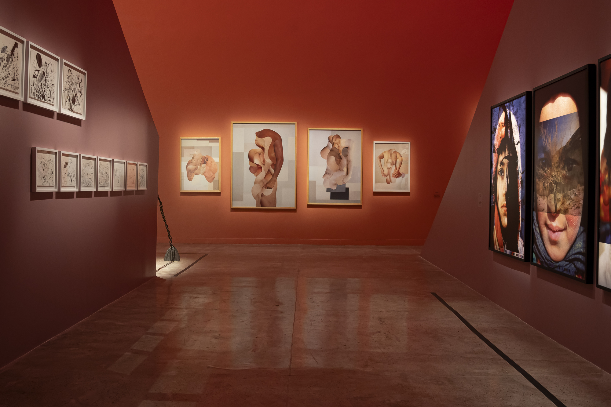 Maroon wall of small framed drawings beside a back wall of collage nudes and large framed portraits in warm gallery light