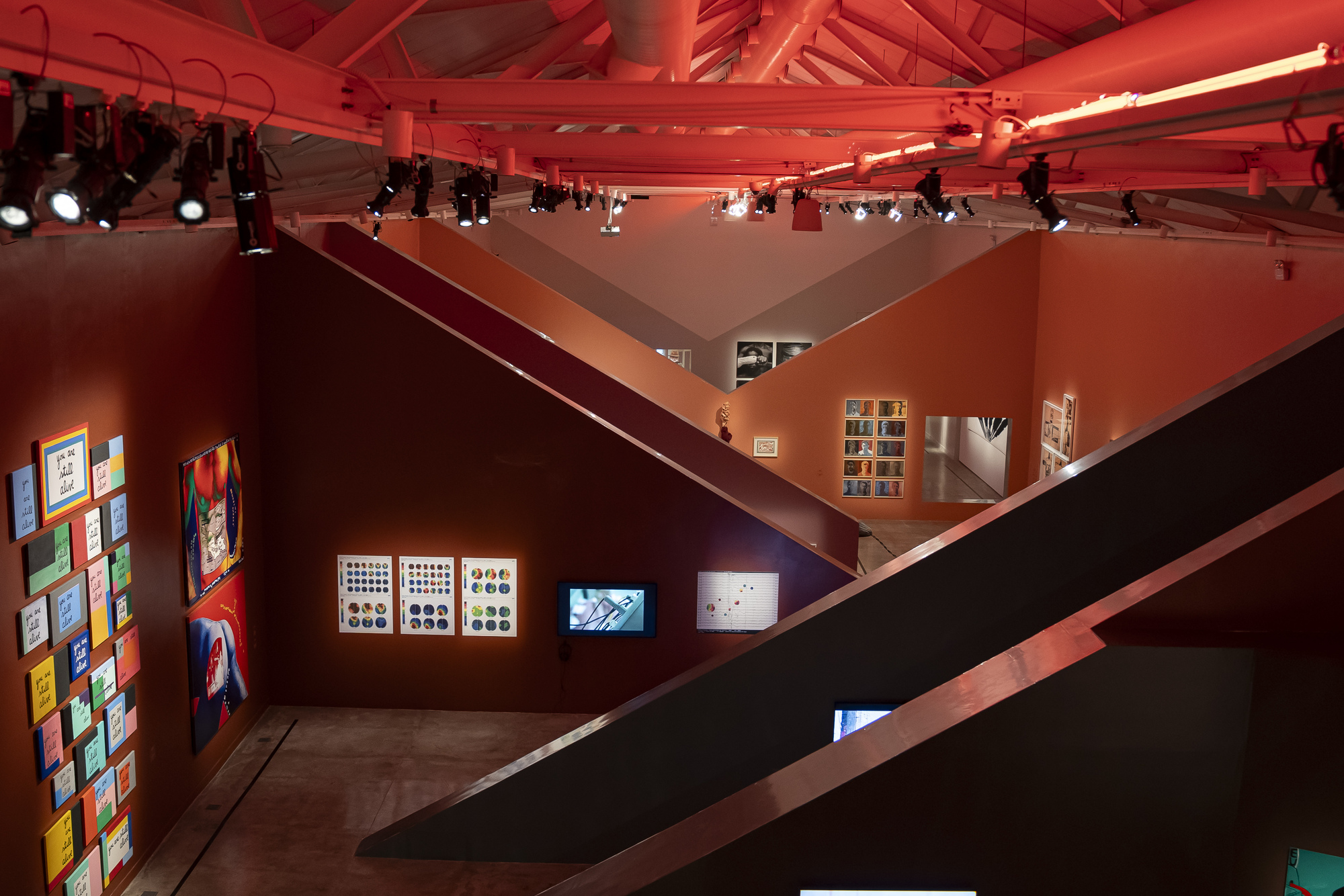 High view over sloping partition walls with track spotlights, tiled colorful works on the left and monitors in the lower gallery