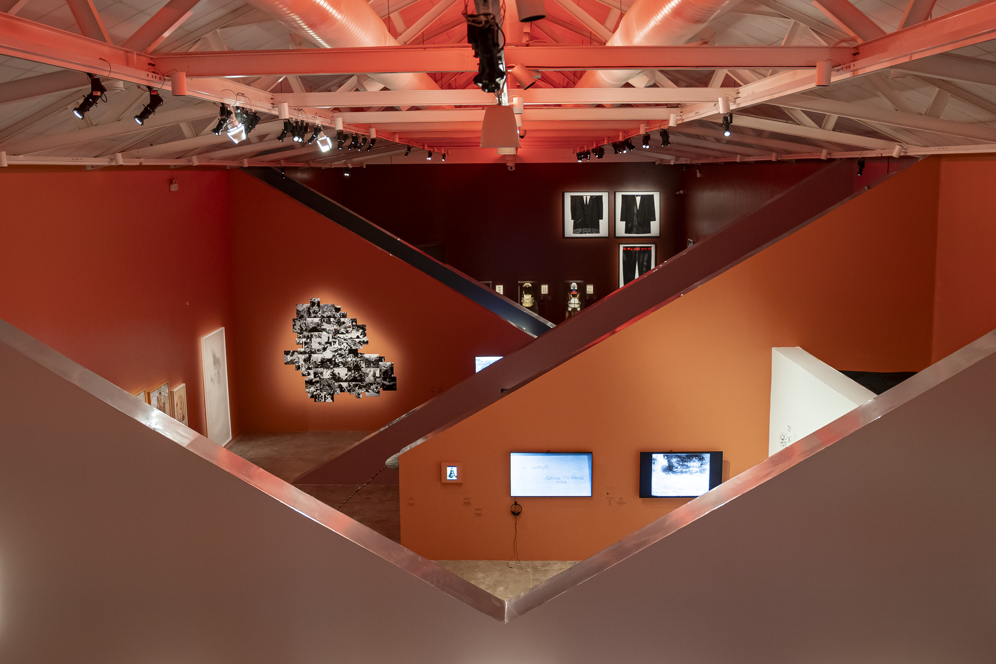 Elevated view through angled walls toward framed black garments, a photo collage and two screens under a red-tinted truss roof