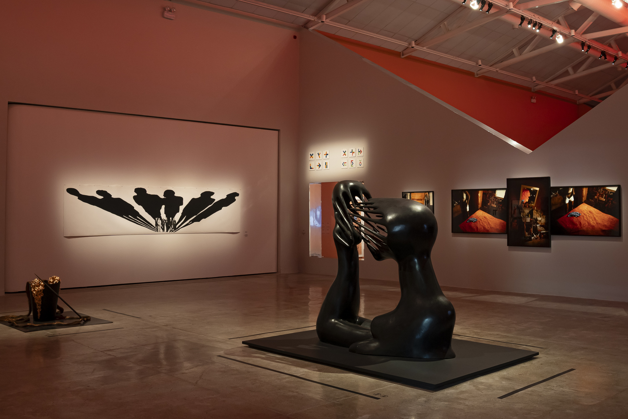 Gallery hall with a black abstract sculpture on a low plinth, a shadow-figure print and lit photographs under warm spotlights