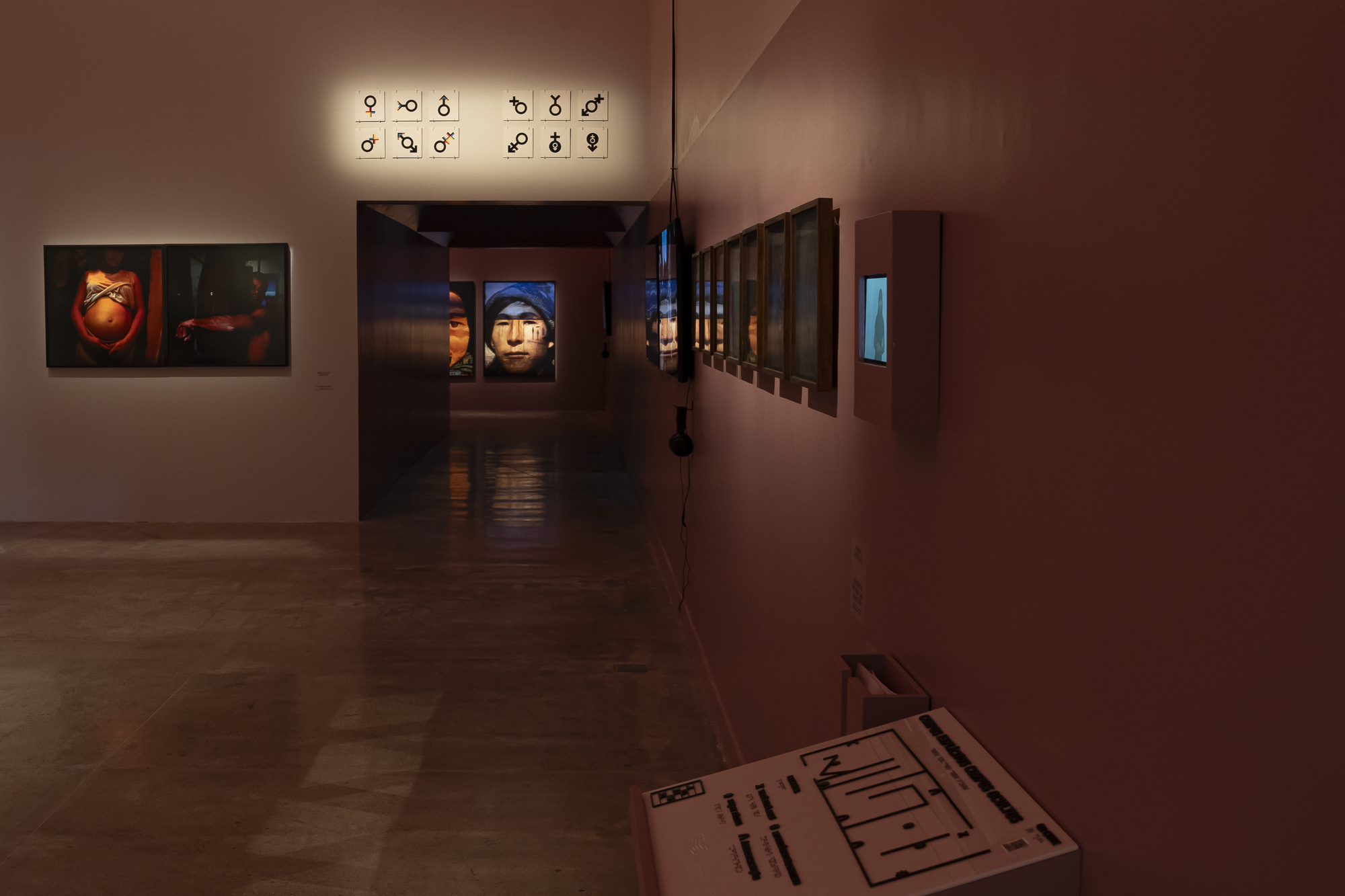Dim corridor of framed portraits and a symbol grid, with a tactile floor-plan model in the foreground