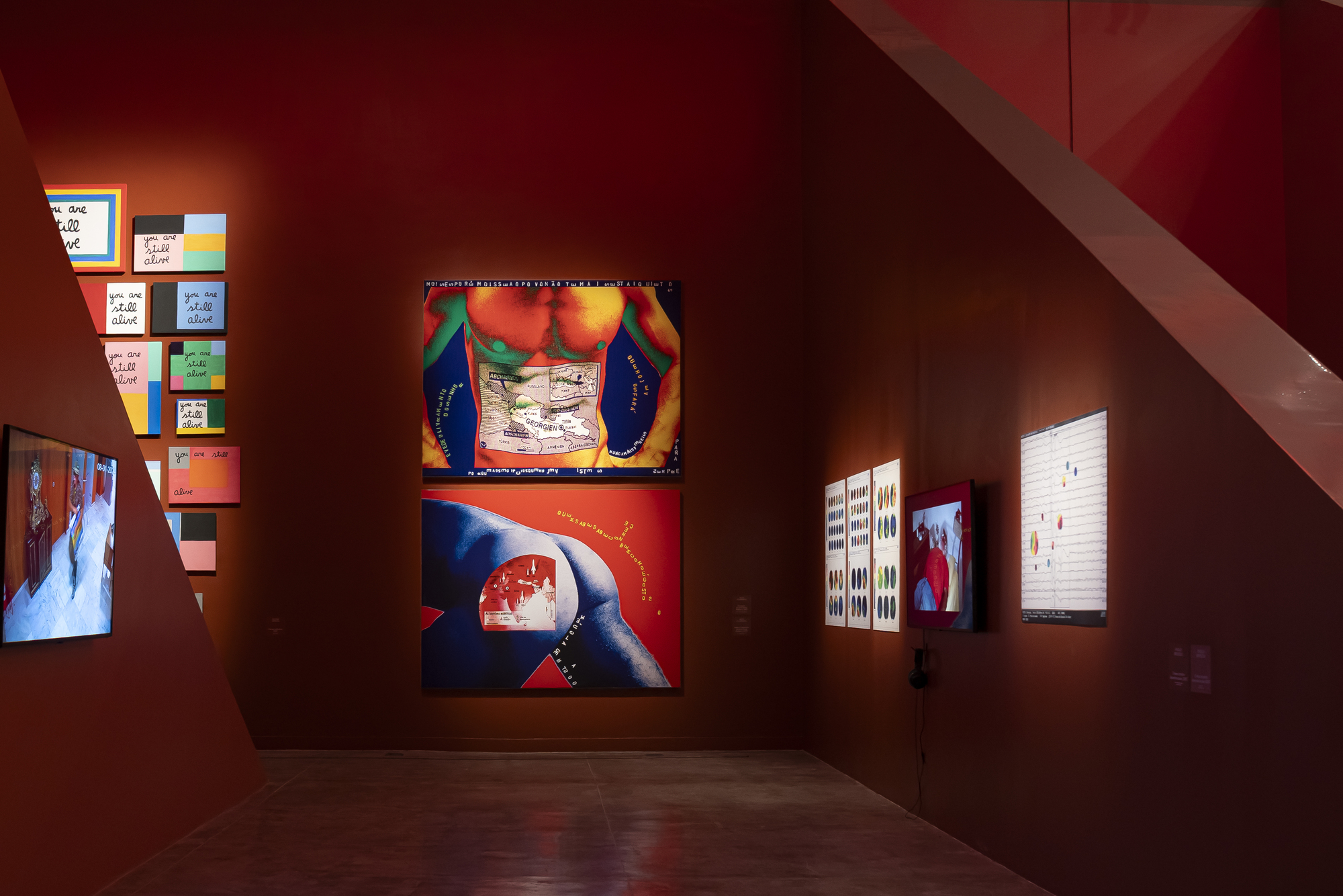 Red-walled room hung with colorful painted panels, a large body artwork and small screens beside an angled partition