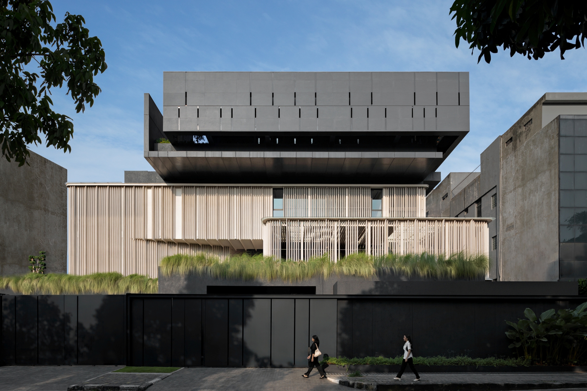 Street facade of stacked dark metal-clad boxes above a timber-fin screen and grass-planted terrace, with two pedestrians walking past in late light