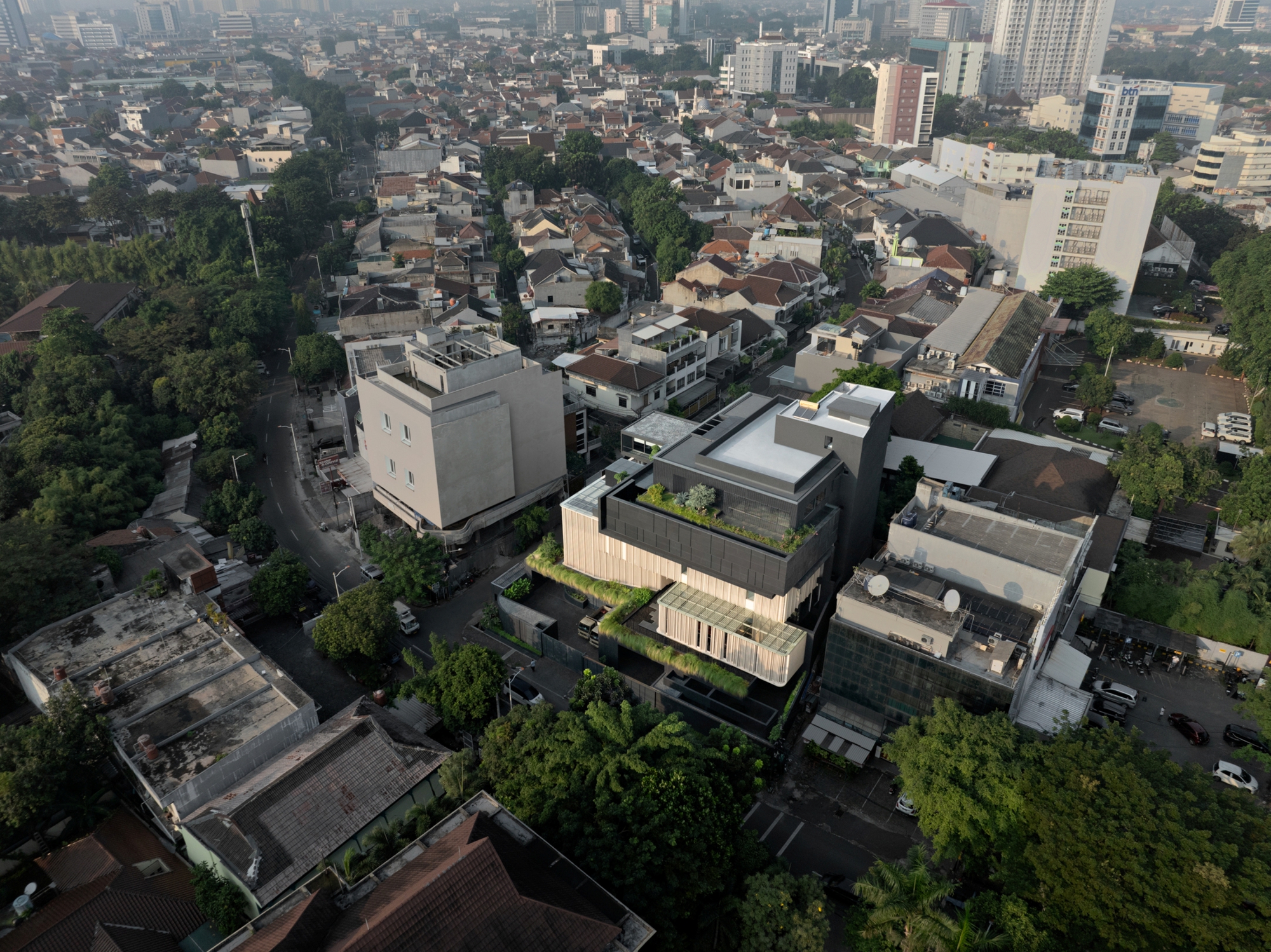 Aerial view of the stacked building with rooftop planting among low houses and dense tree canopy in morning haze