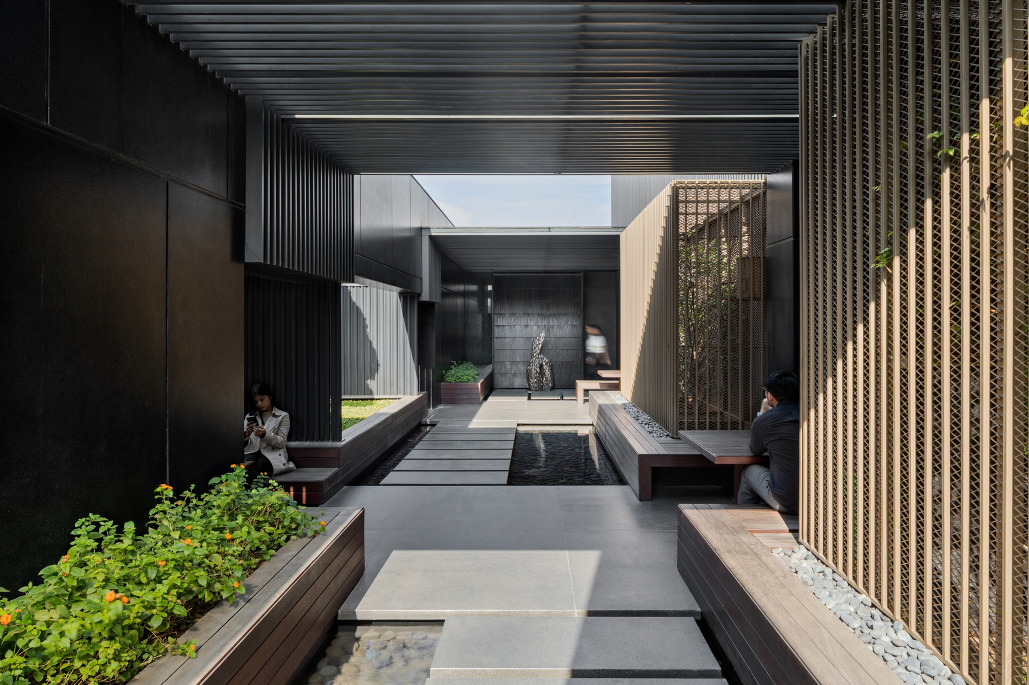 Courtyard passage with stepping-stone slabs over a shallow pool, timber benches, flowering planters and seated people between dark panels and vertical slats