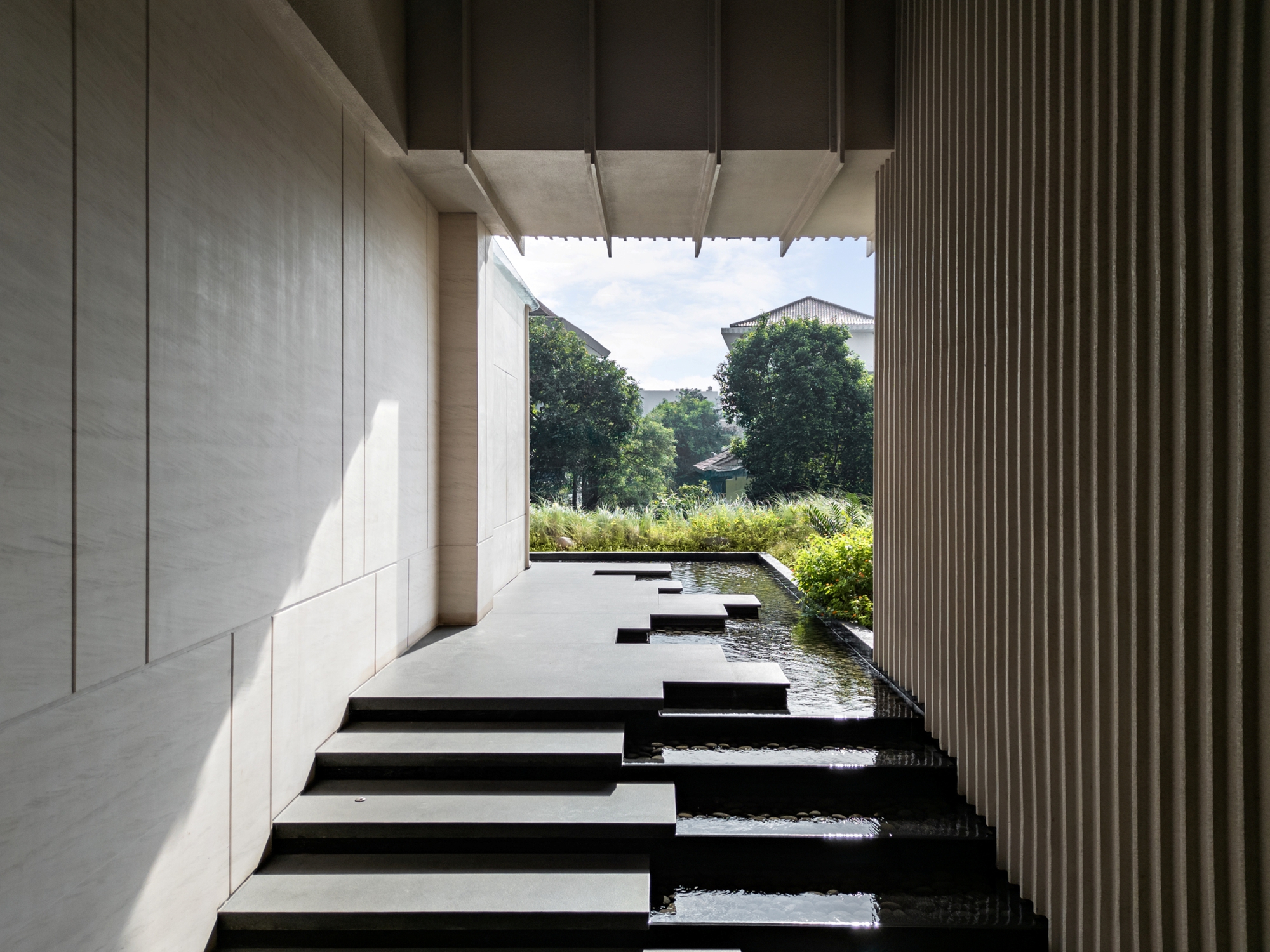 Covered walkway with black stone steps descending into a narrow water channel, framed by stone cladding and fluted wall toward green reeds