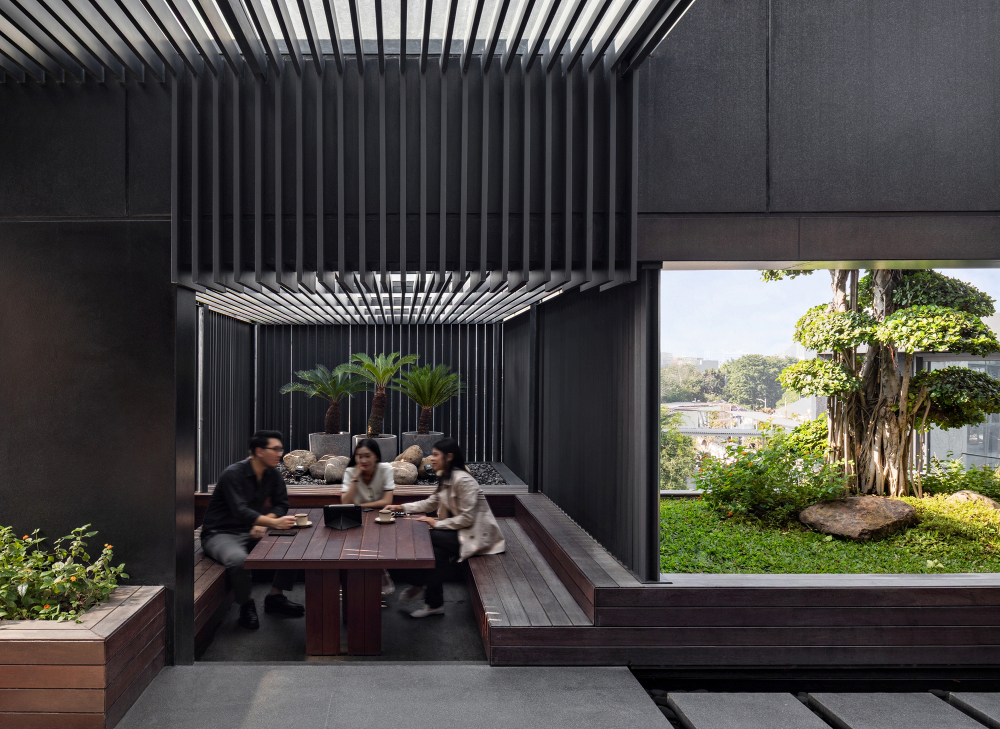 Sunken timber seating nook under a louvered canopy with people at a table, beside a rooftop garden with a large banyan-like tree