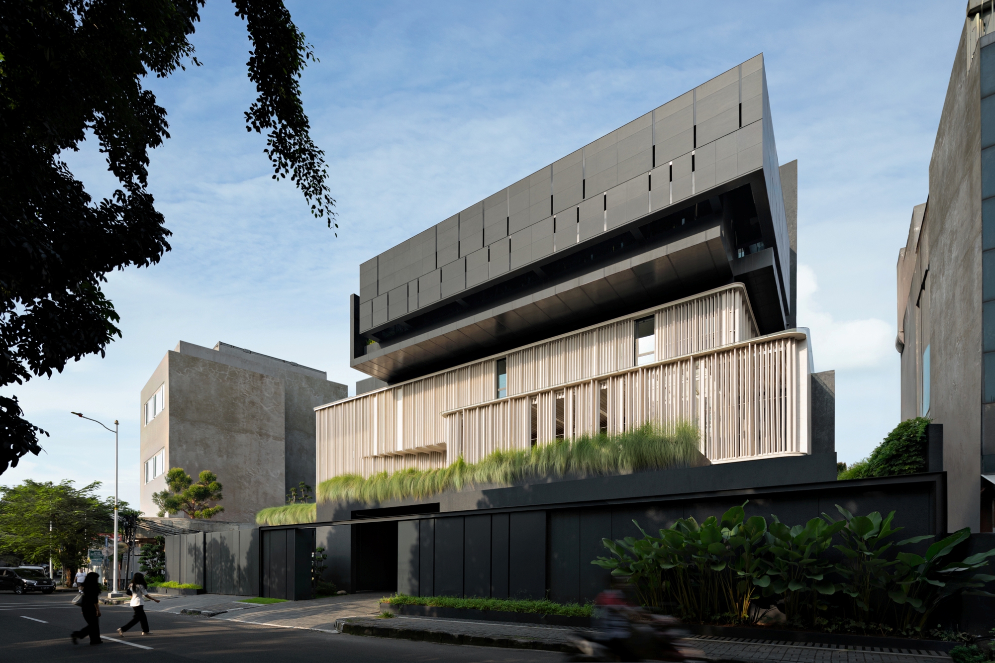 Street view of the stepped facade with timber-toned vertical fins over a dark base, grass planting and a pedestrian crossing at dusk