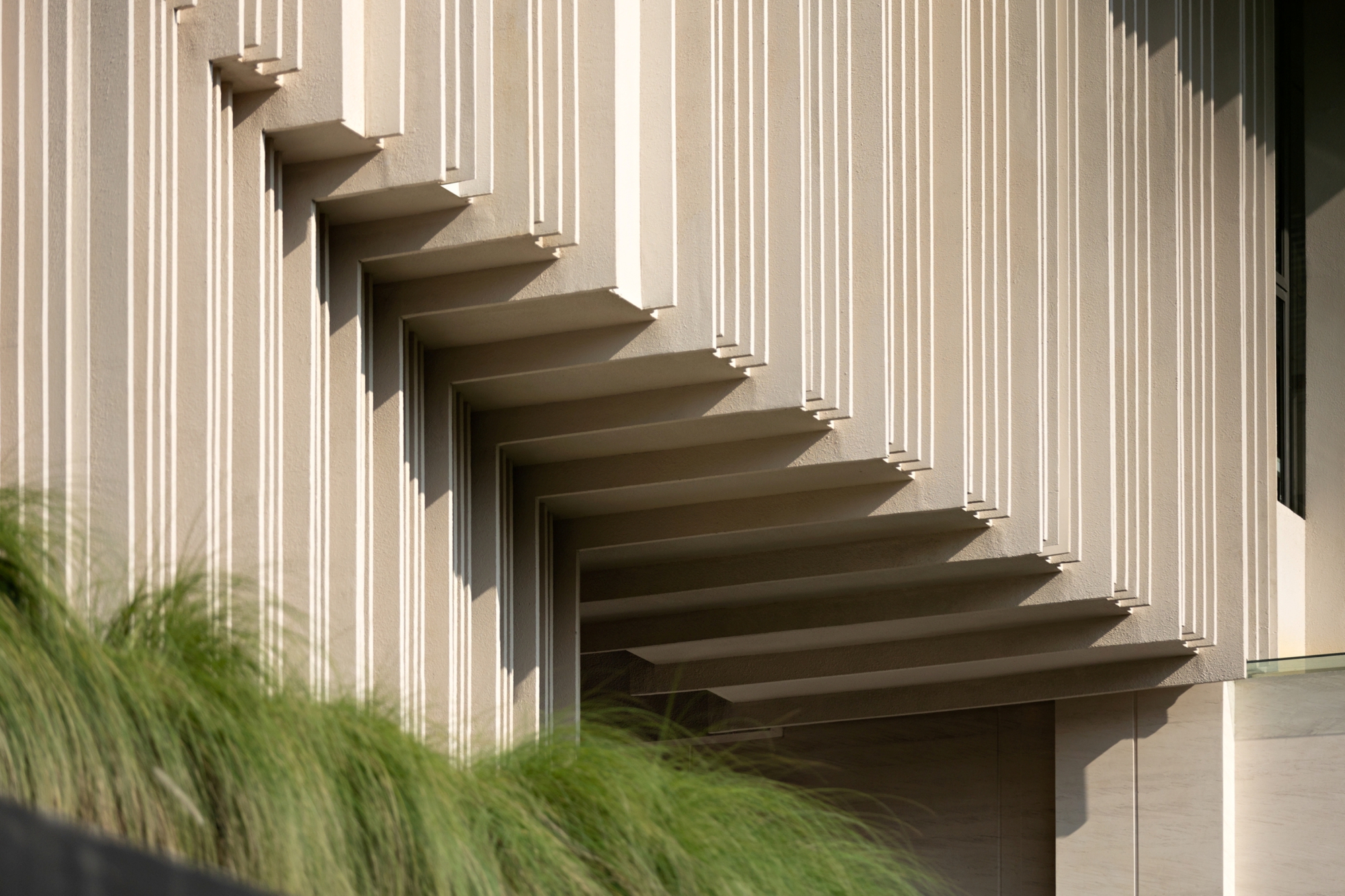 Close-up of stepped vertical fins on the pale facade, with shadows falling across them above a clump of feathery grass