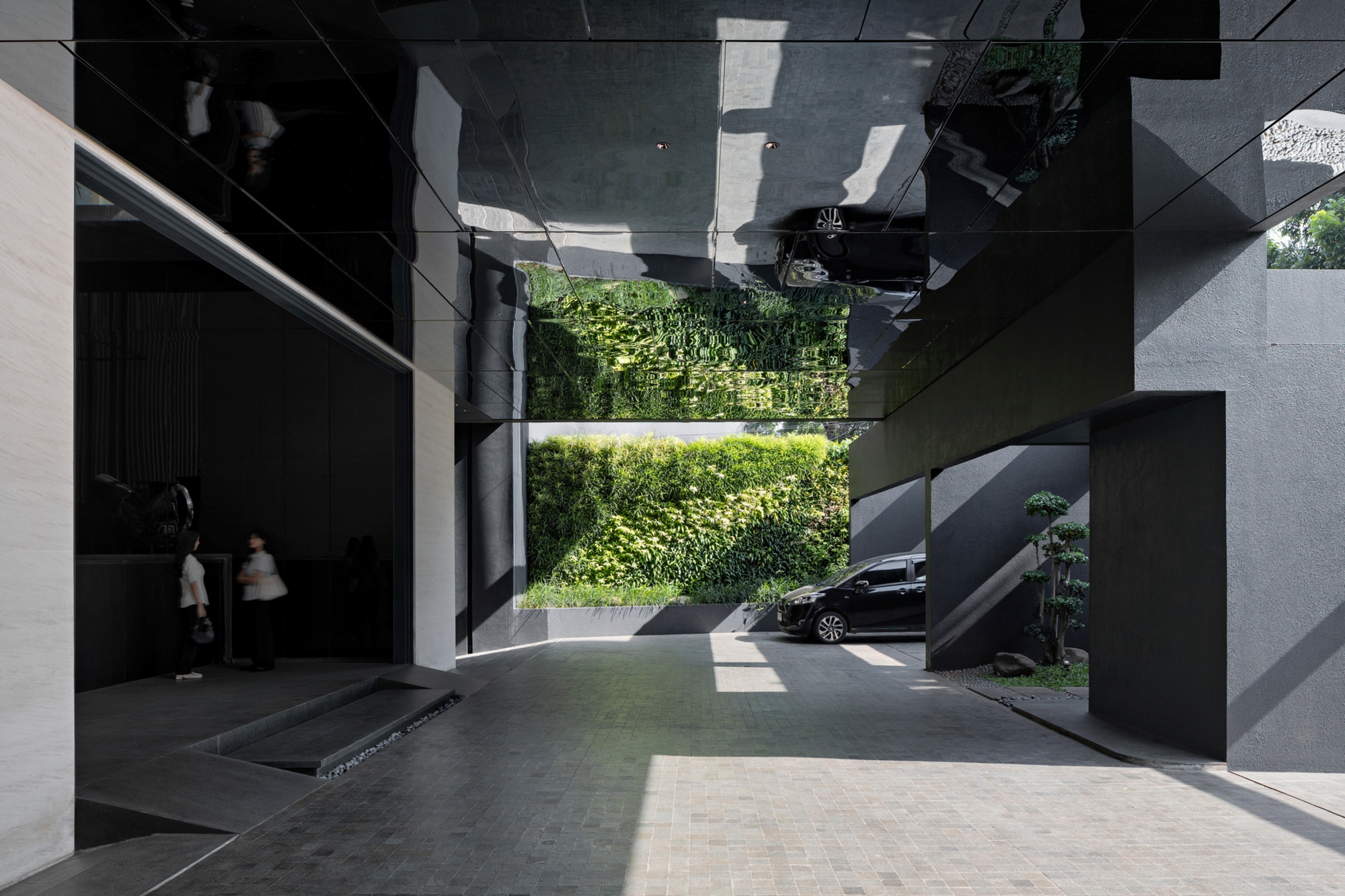 Covered drop-off court with a mirrored black ceiling reflecting a green wall, a parked car and two people near the lobby