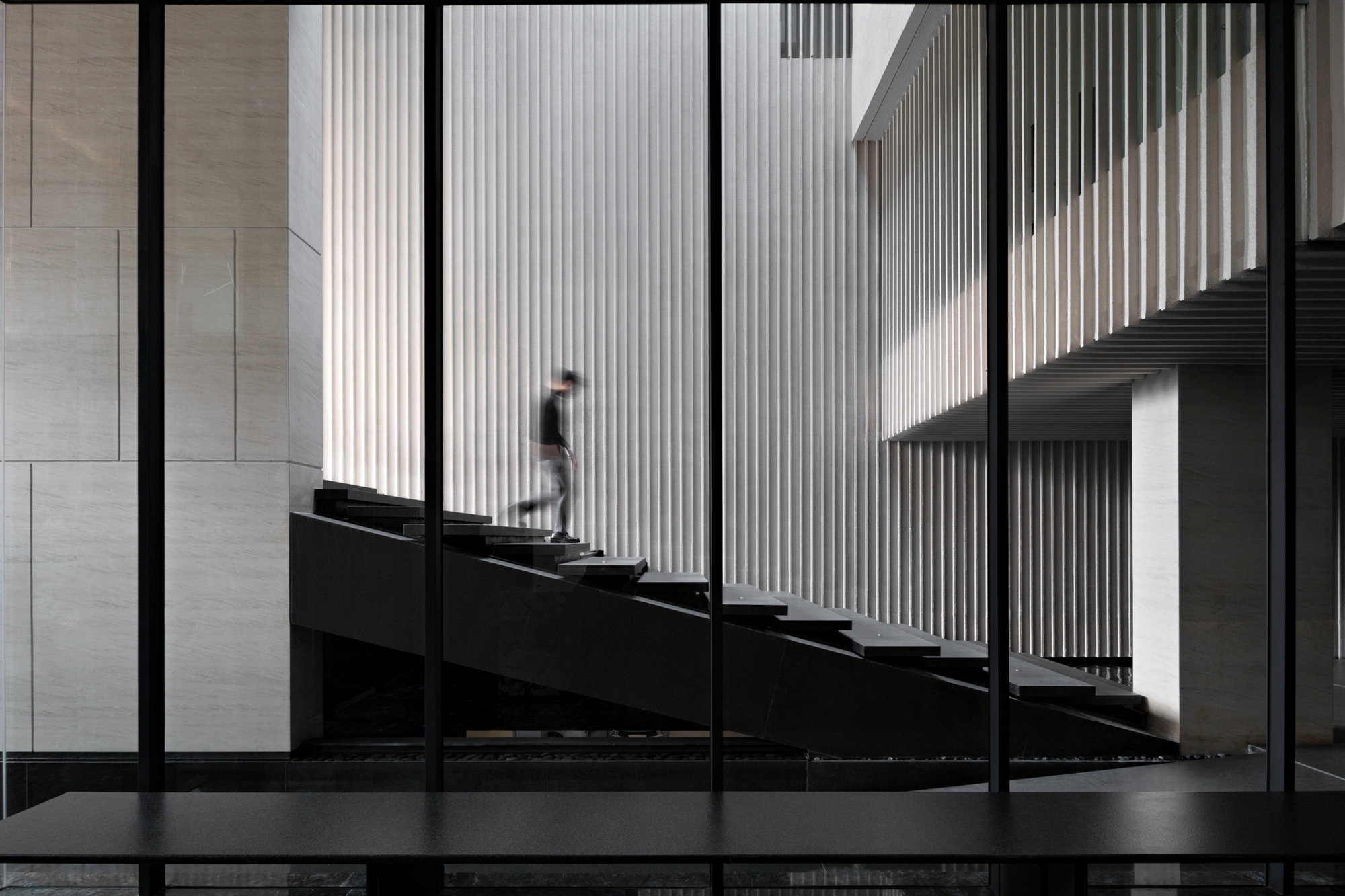 Dark cantilevered stair rising past a fluted white wall, with a blurred figure descending behind slim glazing mullions