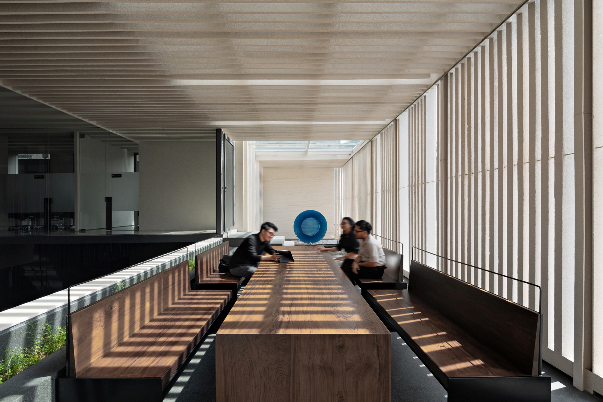 Long timber table with built-in benches under a slatted ceiling, where three people sit in sunlight filtered through vertical fins