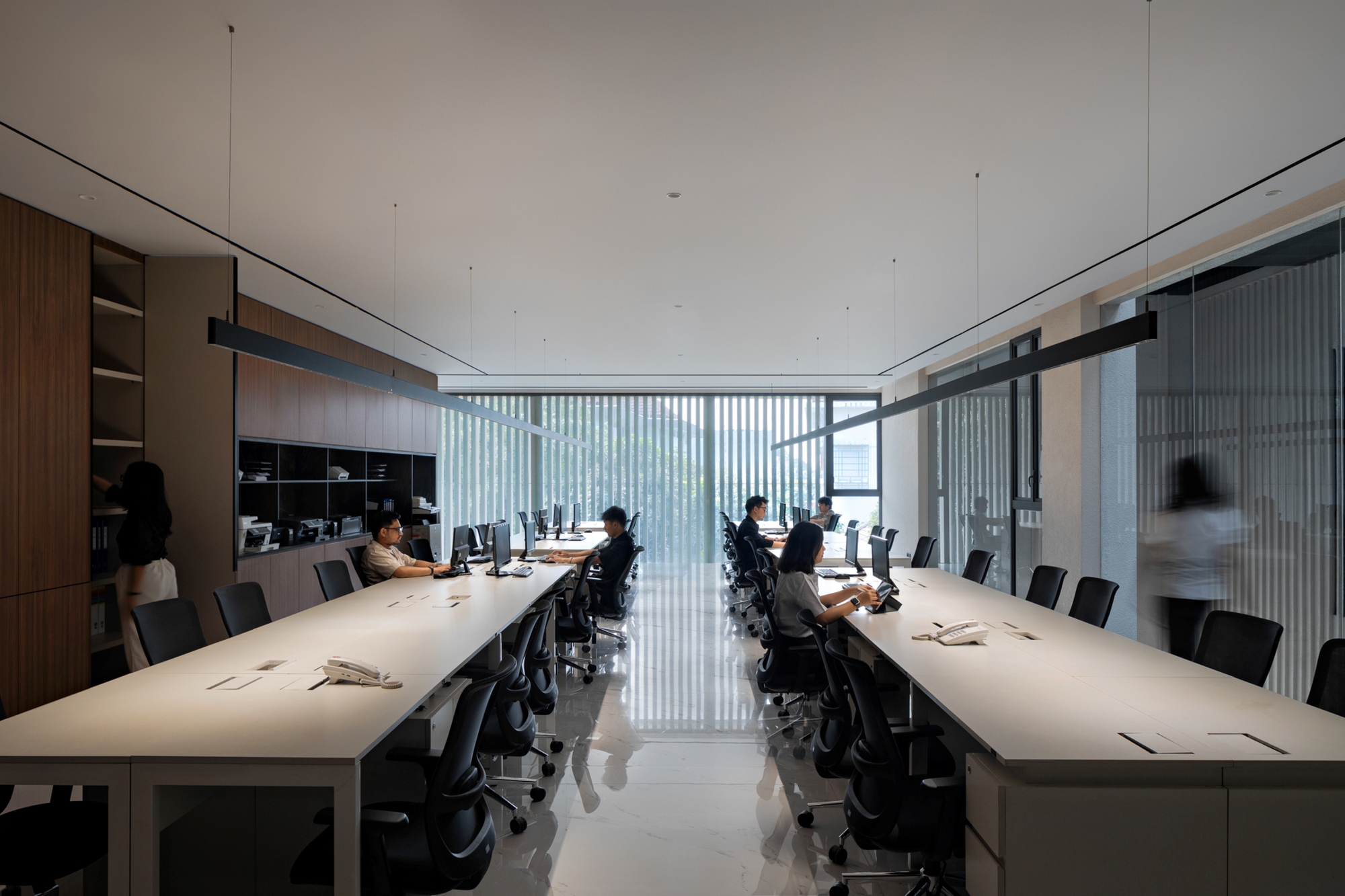 Open office with two long white desks, black task chairs and pendant light bars, facing a sheer-curtained window with blurred staff