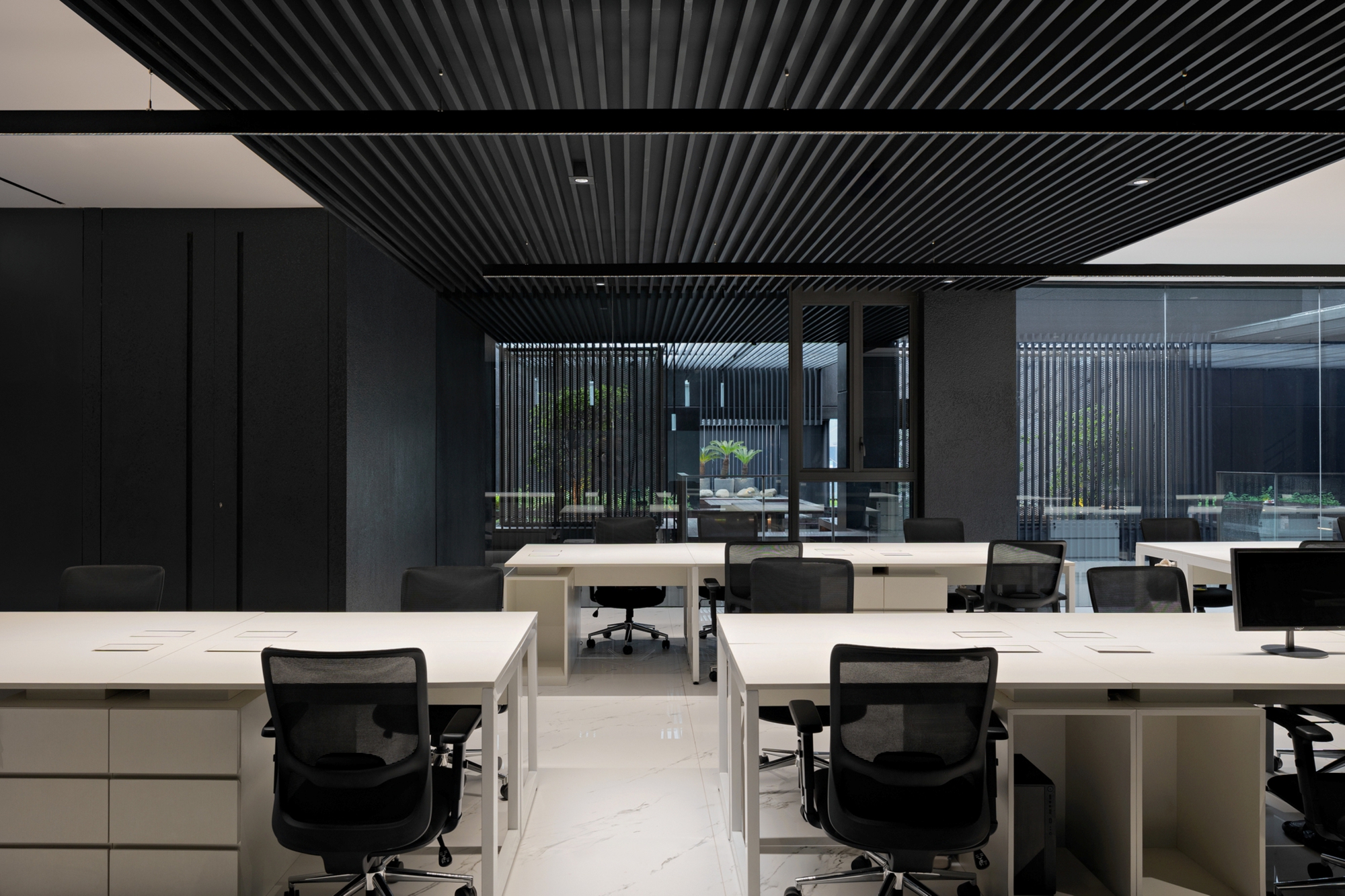 Workstations with mesh chairs beneath a black slatted ceiling, looking through glass to a planted courtyard with vertical screens