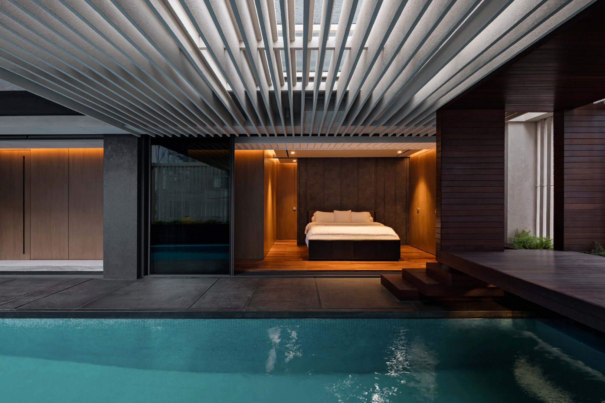 Bedroom opening onto a lap pool beneath a fin-slatted canopy, with timber steps and a dark deck at dusk