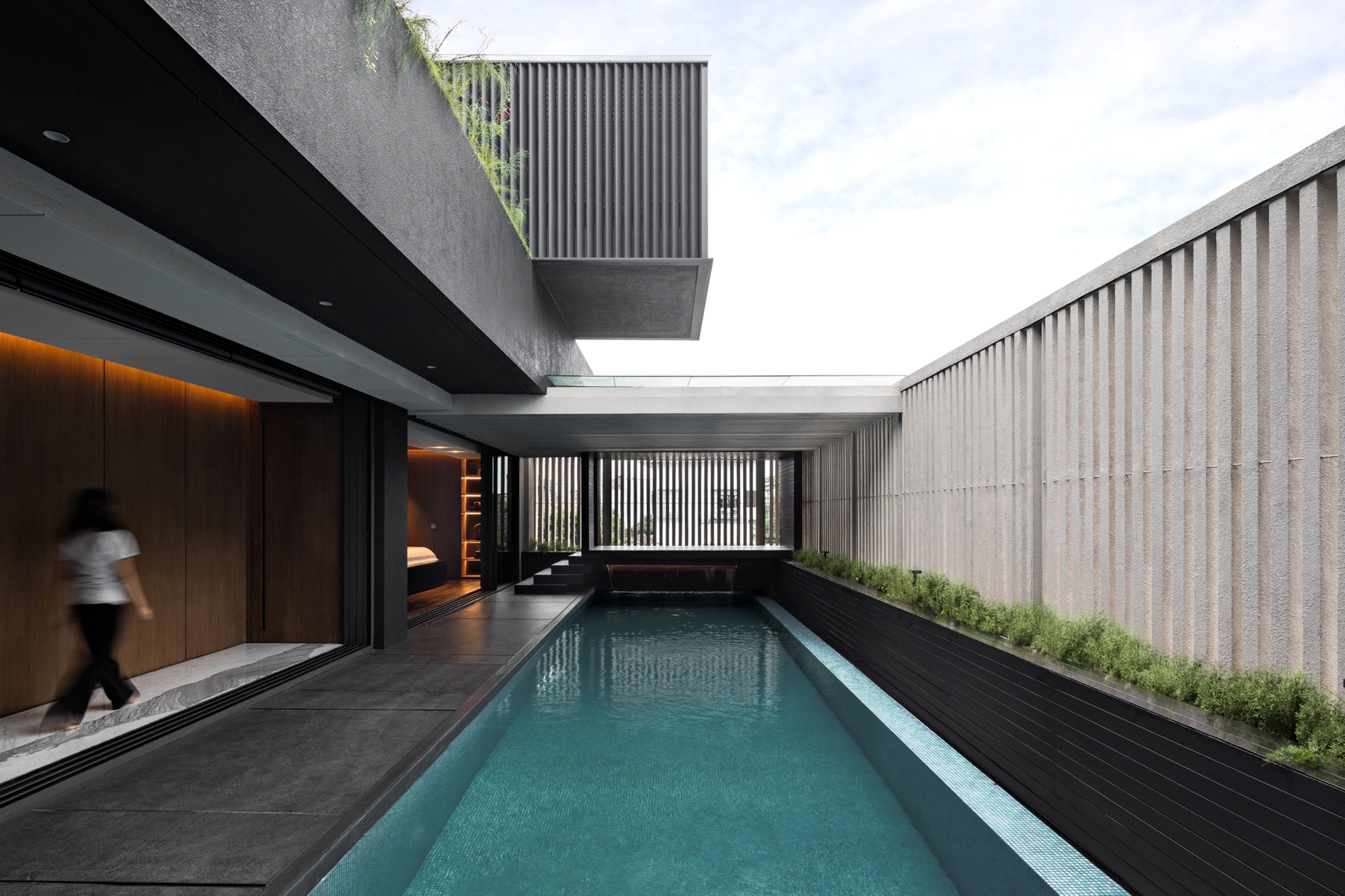 Long narrow pool between a dark cantilevered volume and a ribbed concrete wall, with a woman walking past the glazing