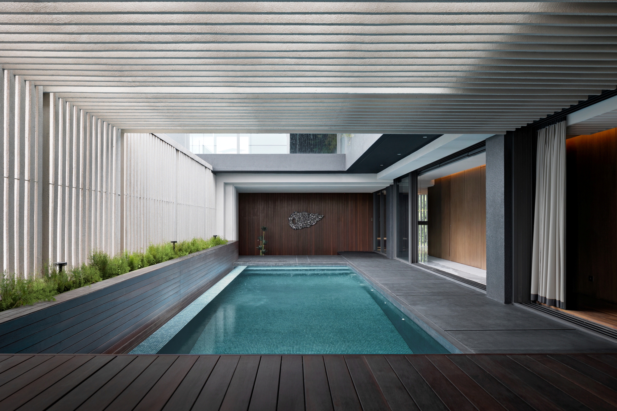 Lap pool framed by a slatted ceiling, ribbed screen wall and planted edge, with a metal wall sculpture at the far end